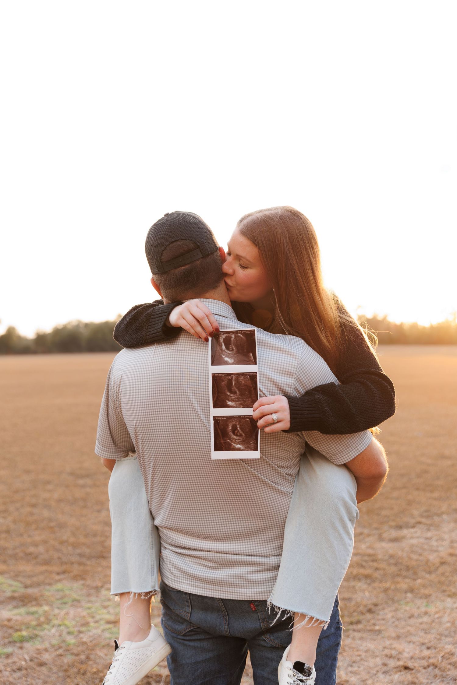 Couple embraces while holding ultrasound photos during sunset in an open field.