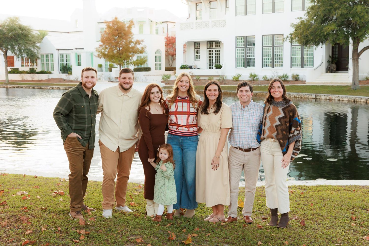 Group of people standing together on grass by a lake with white building in background.