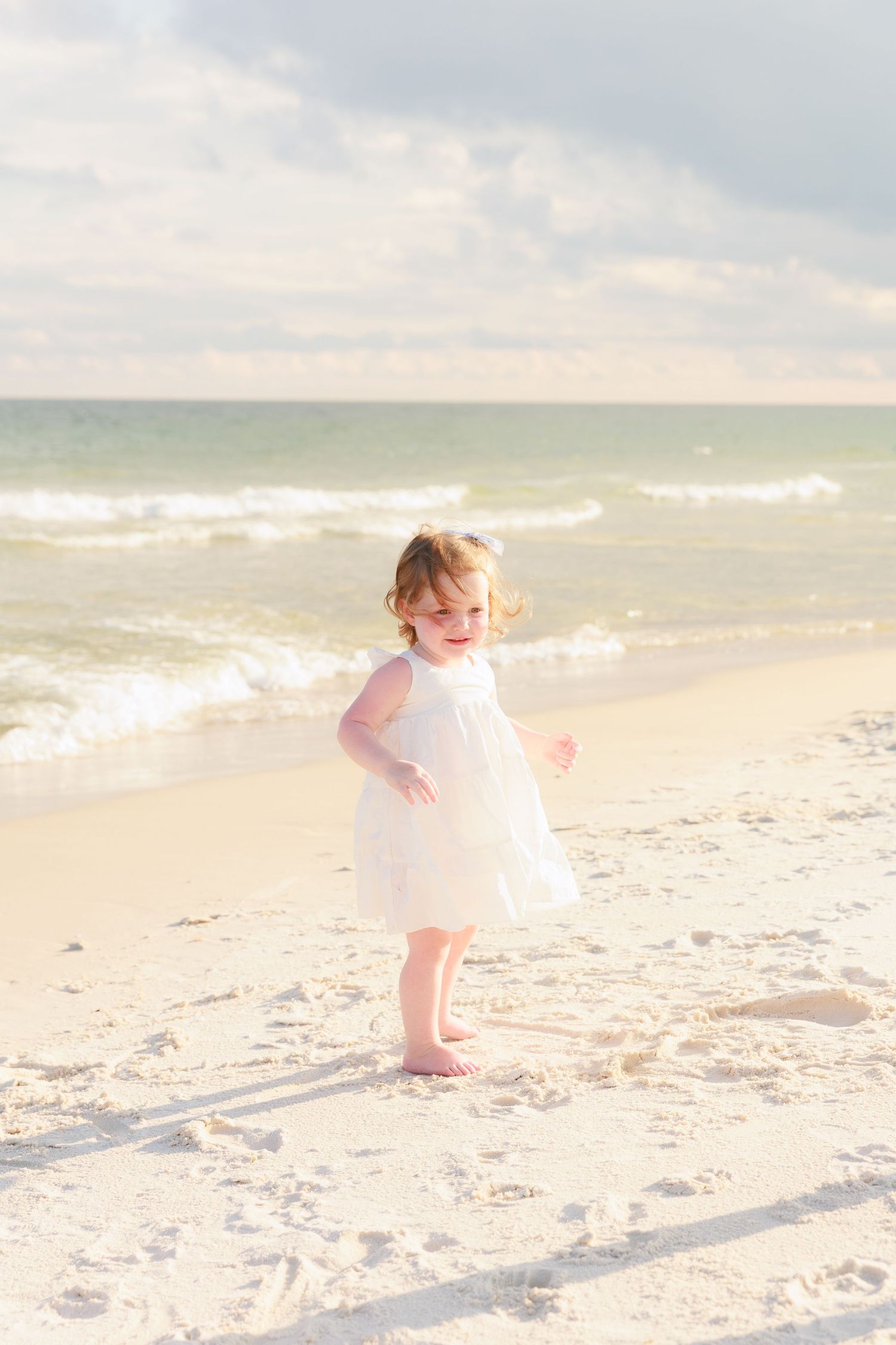 Toddler girl in white dress playing barefoot on sandy beach with ocean waves in background.