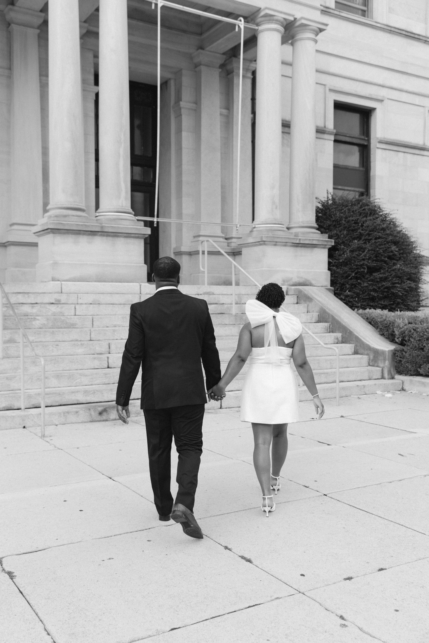 Black and white photo of a couple holding hands walking toward a grand building with columns, dressed in wedding attire.