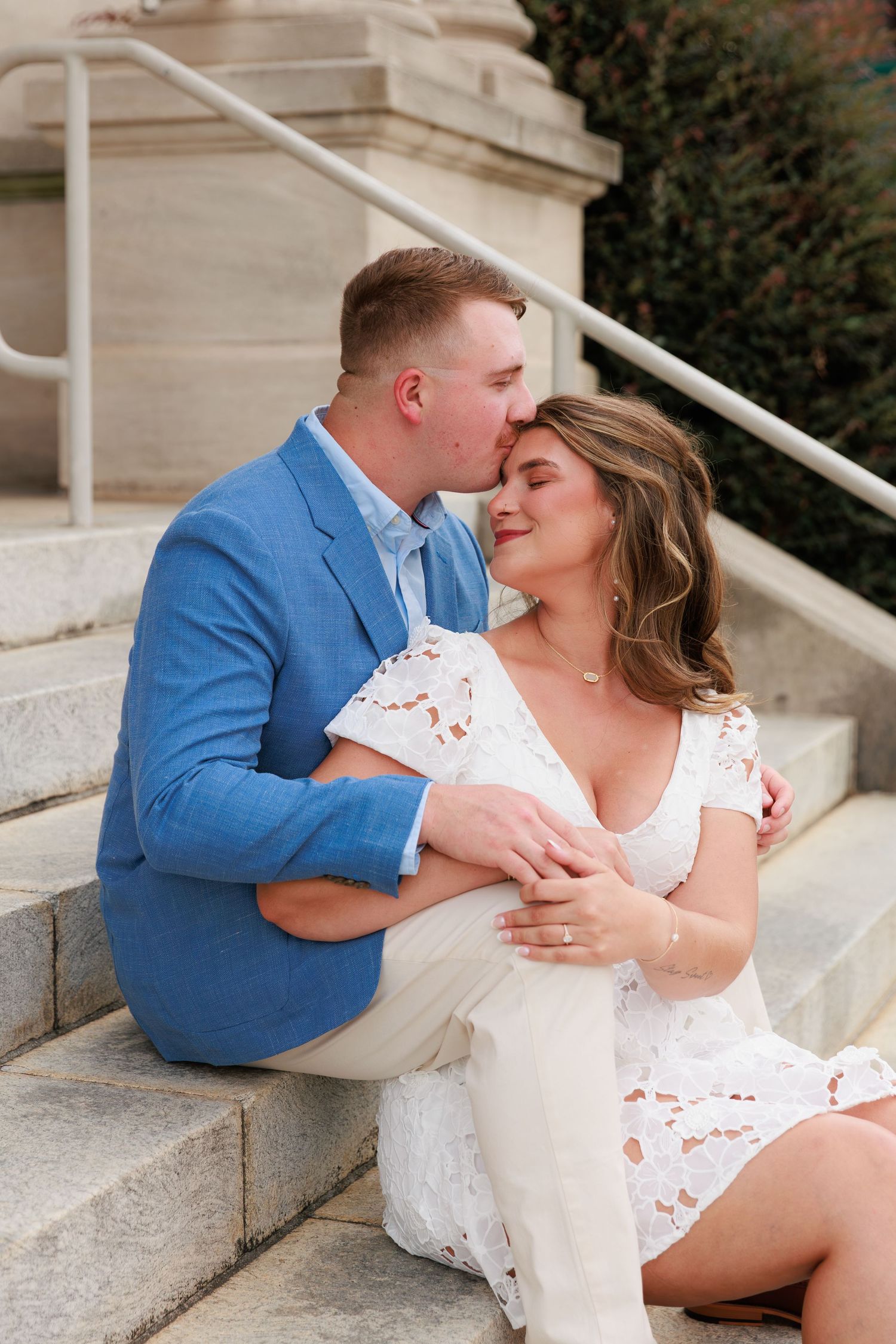A couple sits on stone steps, man in blue blazer kissing woman's forehead as she smiles in white lace dress.