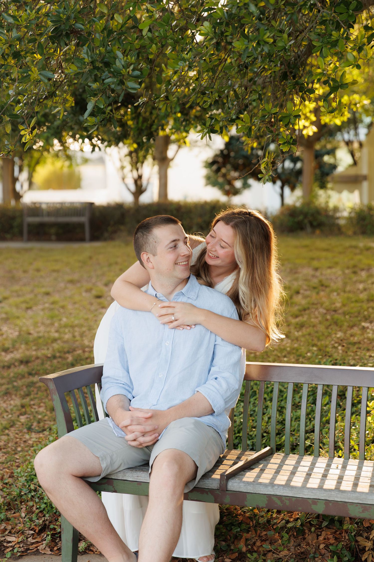 A couple shares a tender moment on a park bench, woman embracing man from behind under lush green trees.