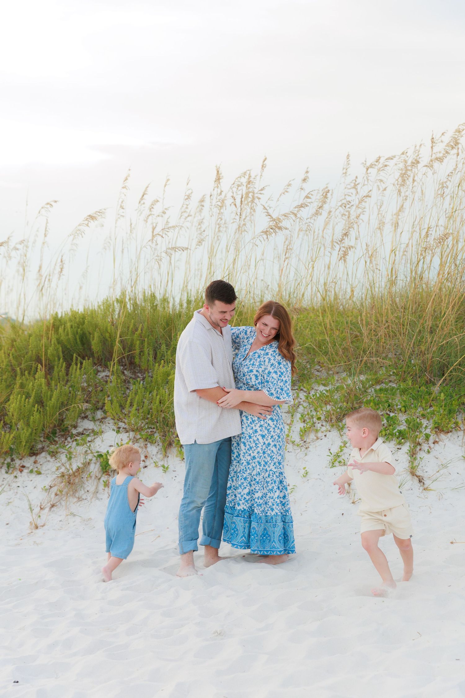Family of four poses on white sand beach dunes, parents embracing while two toddlers play around them.