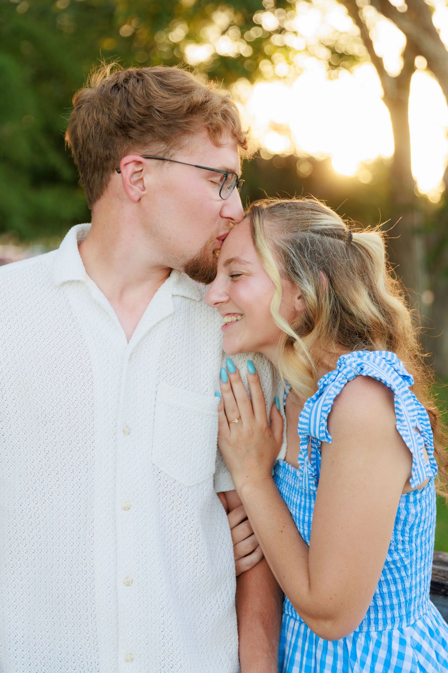A man kisses a smiling woman on the forehead during a romantic outdoor engagement session at golden hour.