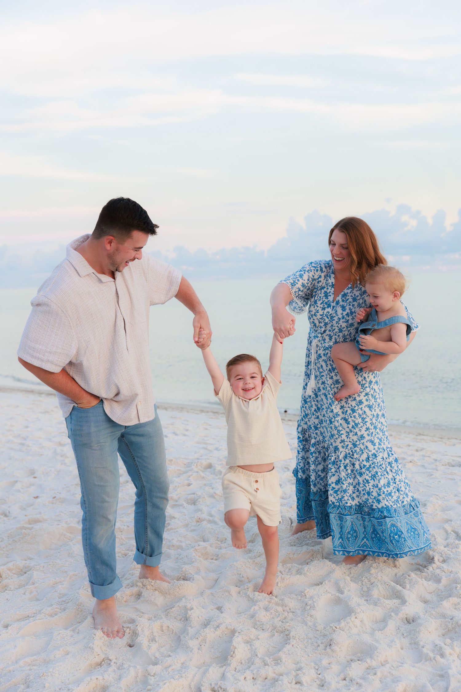 Happy family of four playing on white sand beach at sunset, parents swinging toddler boy while holding baby.