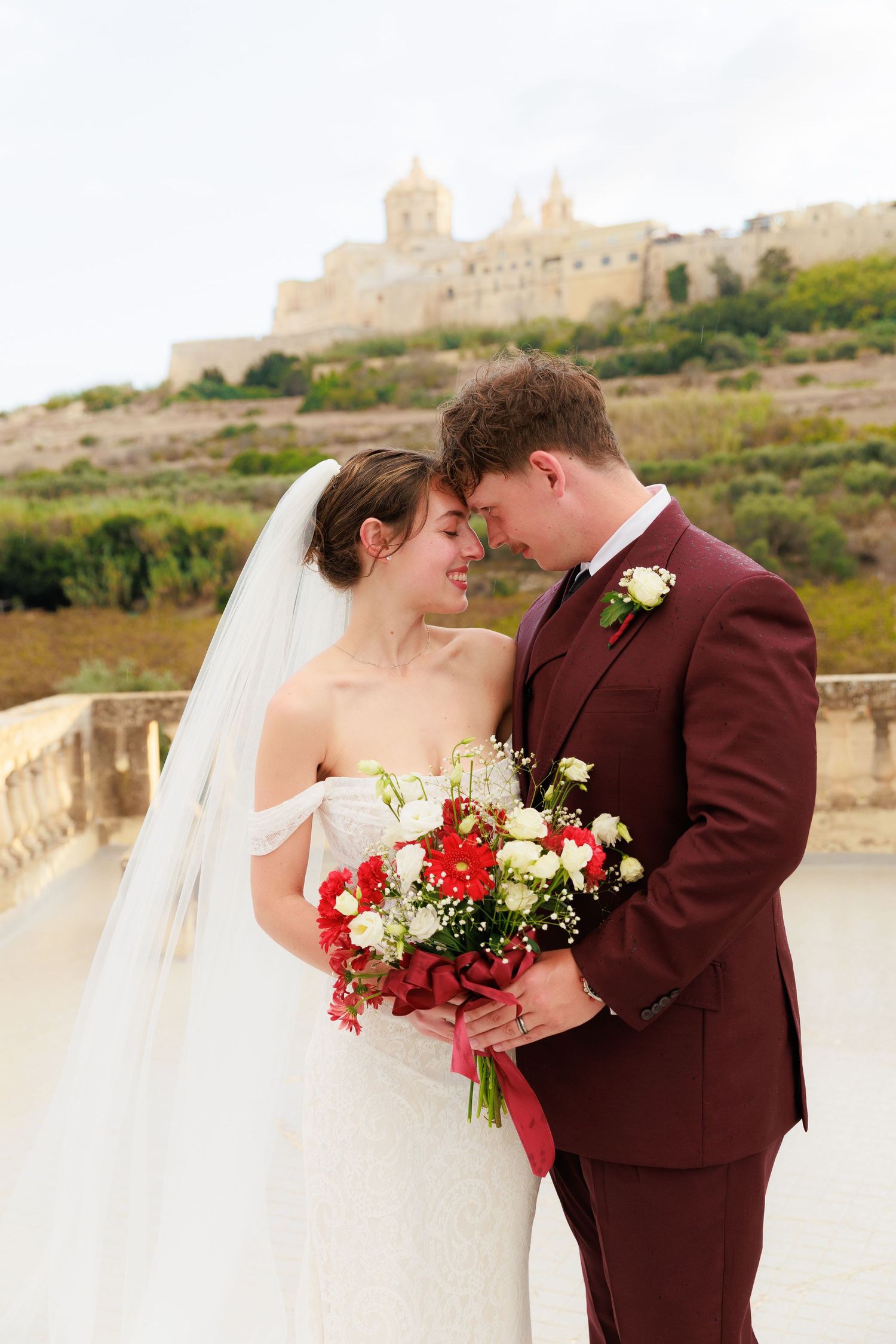 A romantic wedding couple embraces against a scenic stone backdrop while holding a red and white floral bouquet.