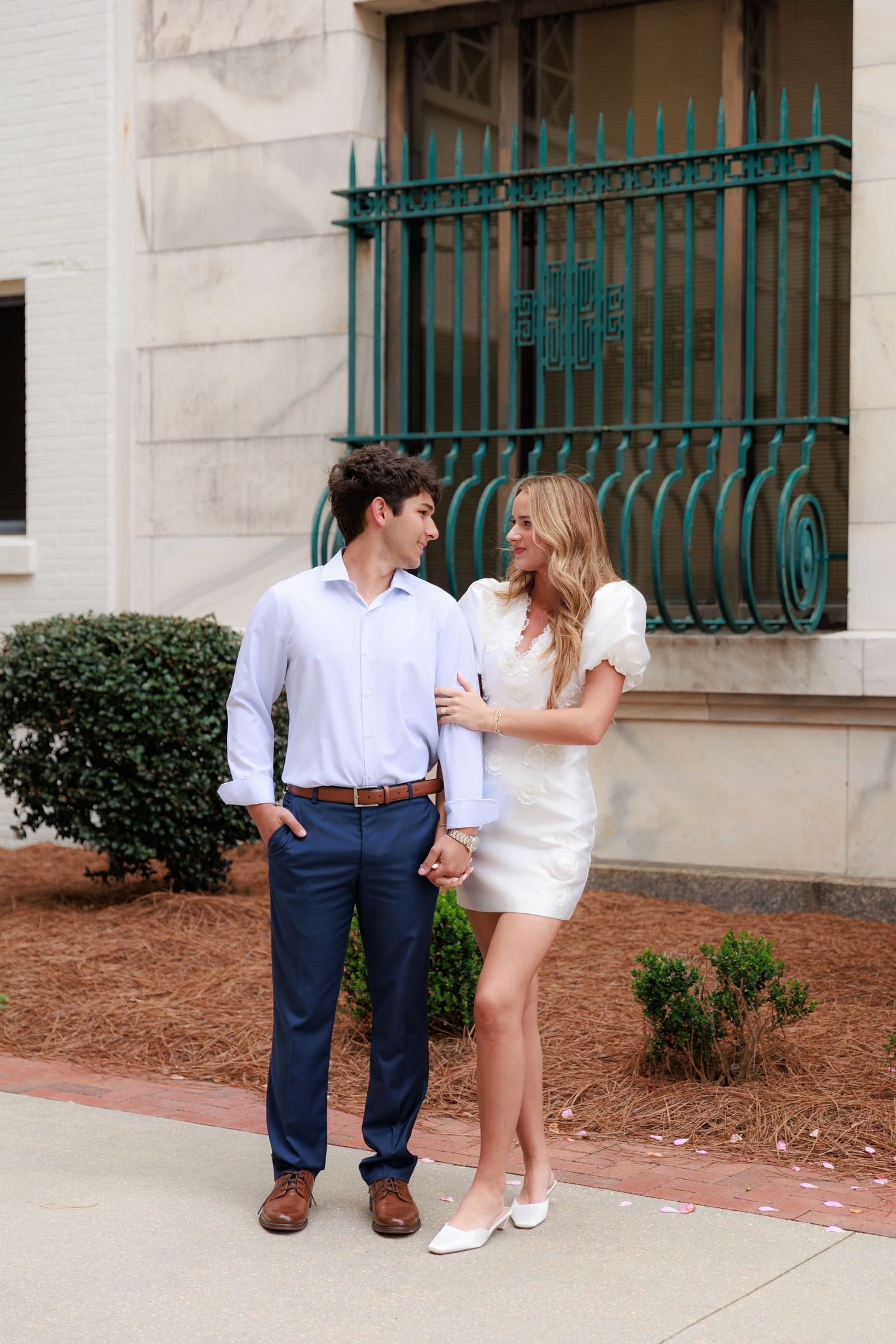 A couple shares a romantic moment on a sidewalk near decorative green railings and manicured shrubs.