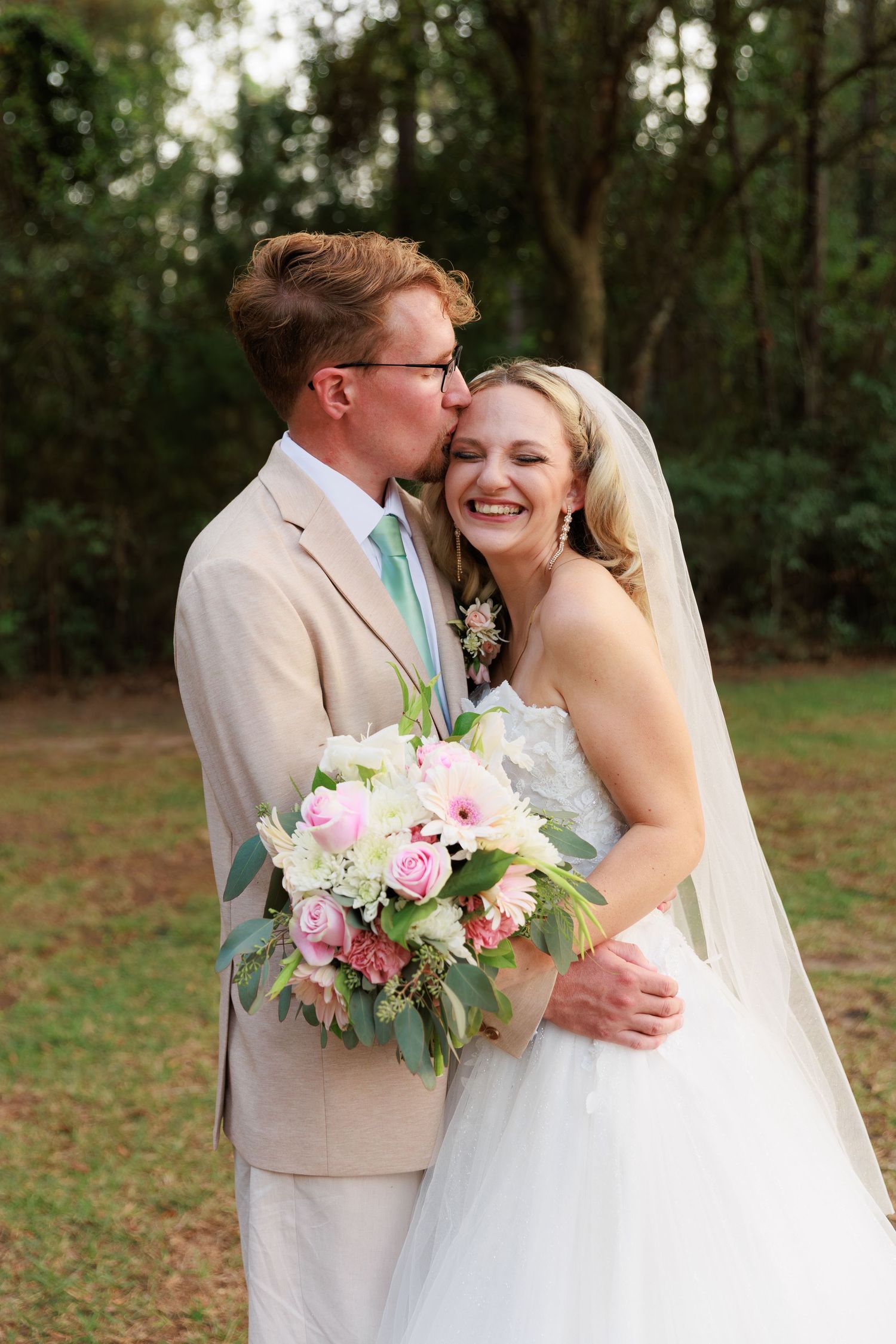 A romantic wedding couple shares joyful moments in a wooded outdoor setting with pink and white floral bouquet.