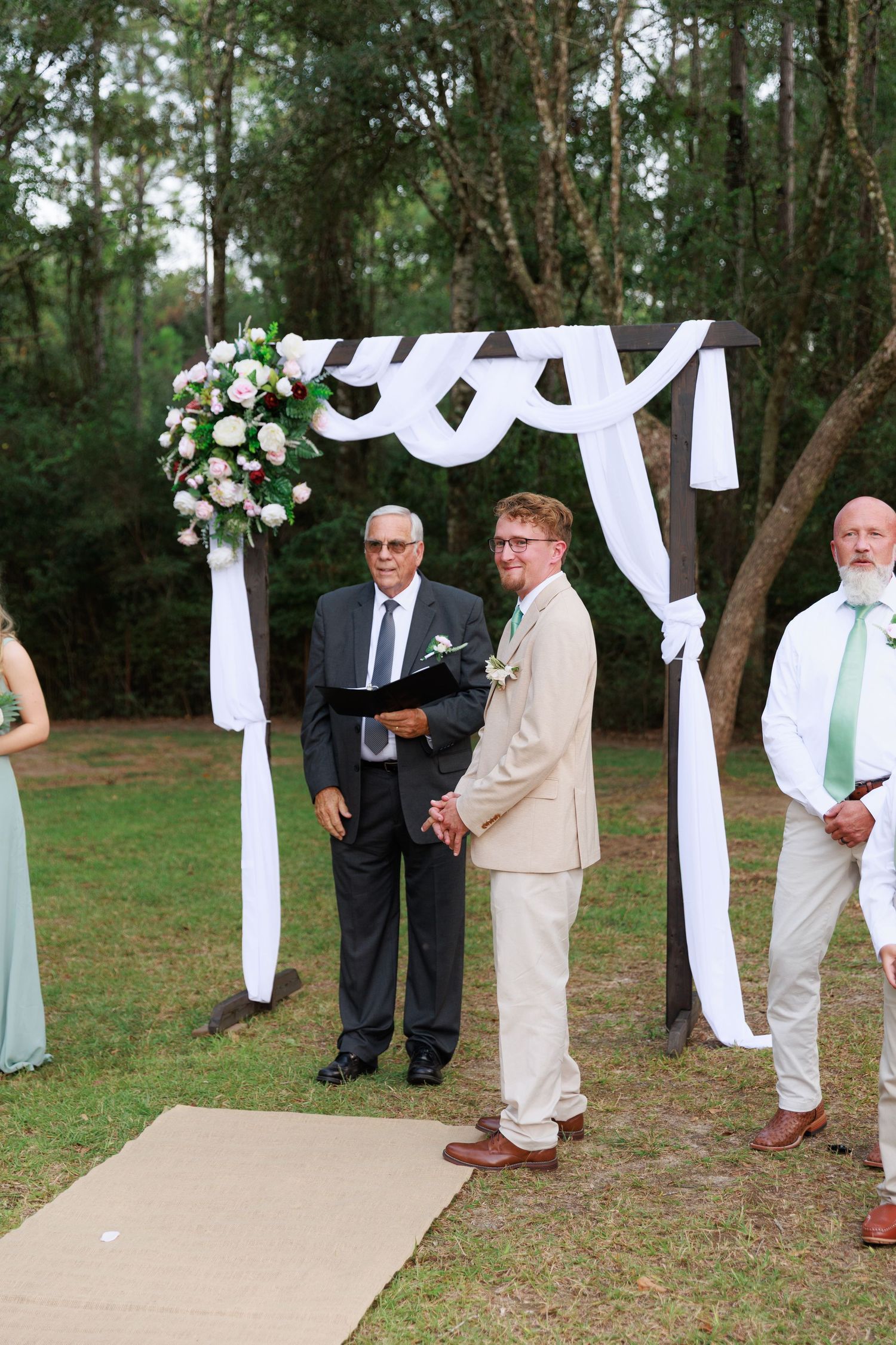 An outdoor wedding ceremony setup with a white draped arch and floral arrangement in a wooded setting.