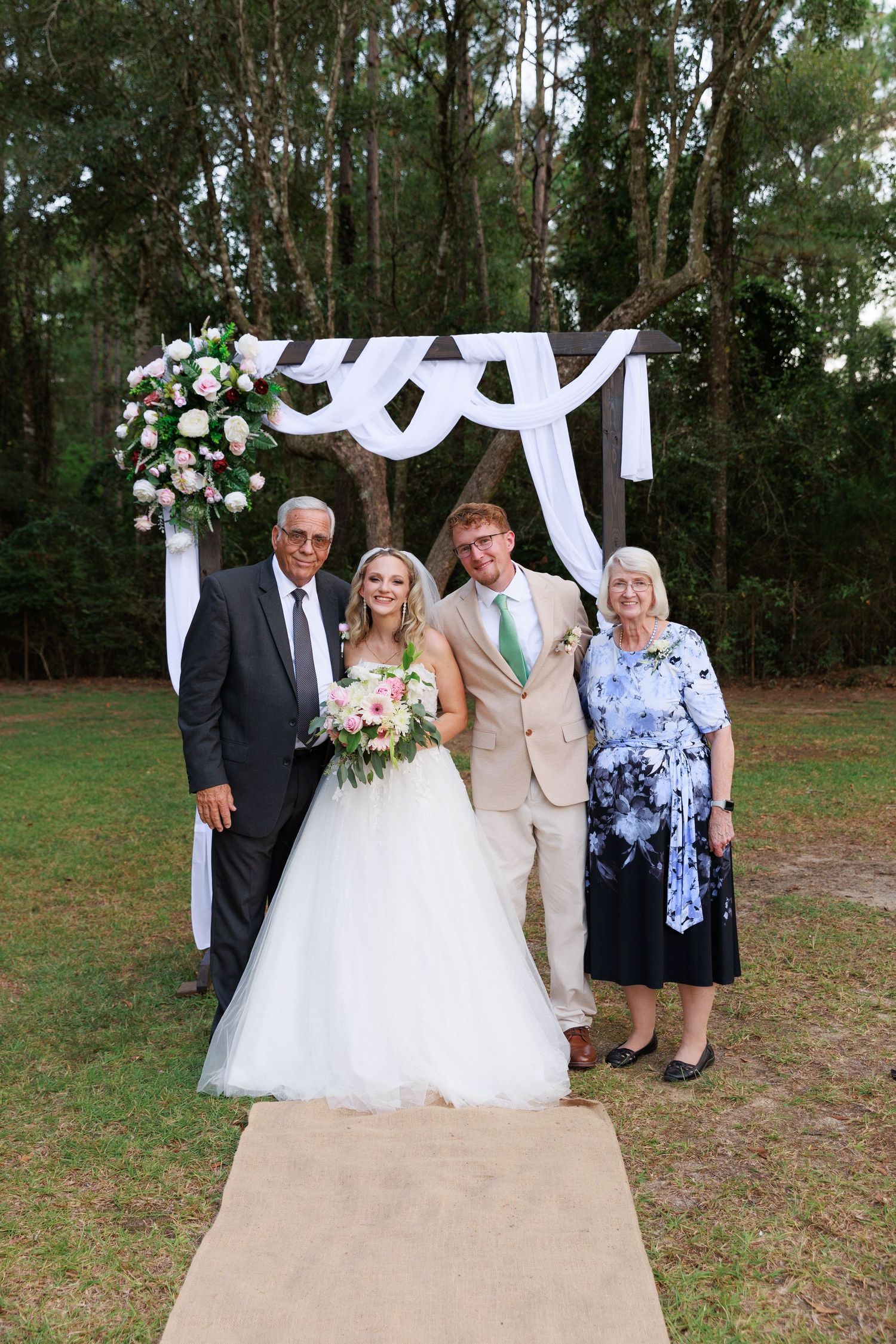 Wedding party members gather under a white draped arch in an outdoor pine forest setting.