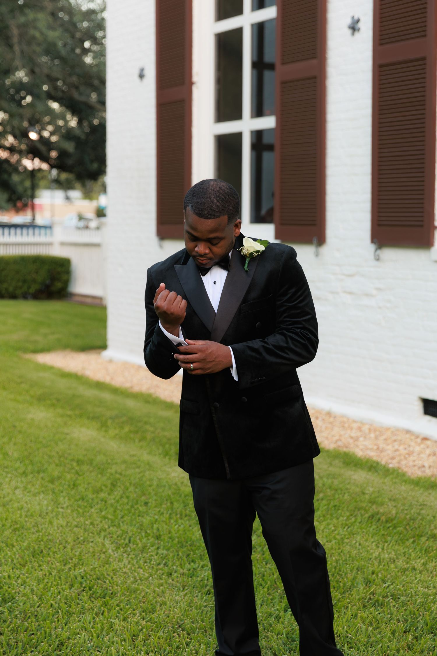 Groom in black tuxedo adjusts his tie outside white church building with brown shutters.
