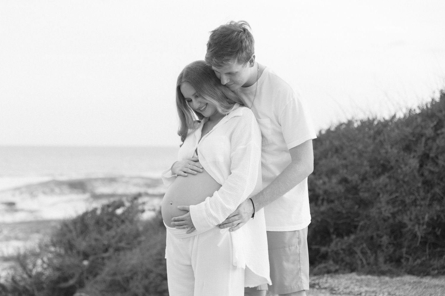 A couple in white outfits embraces on a scenic coastal path during a romantic photoshoot.