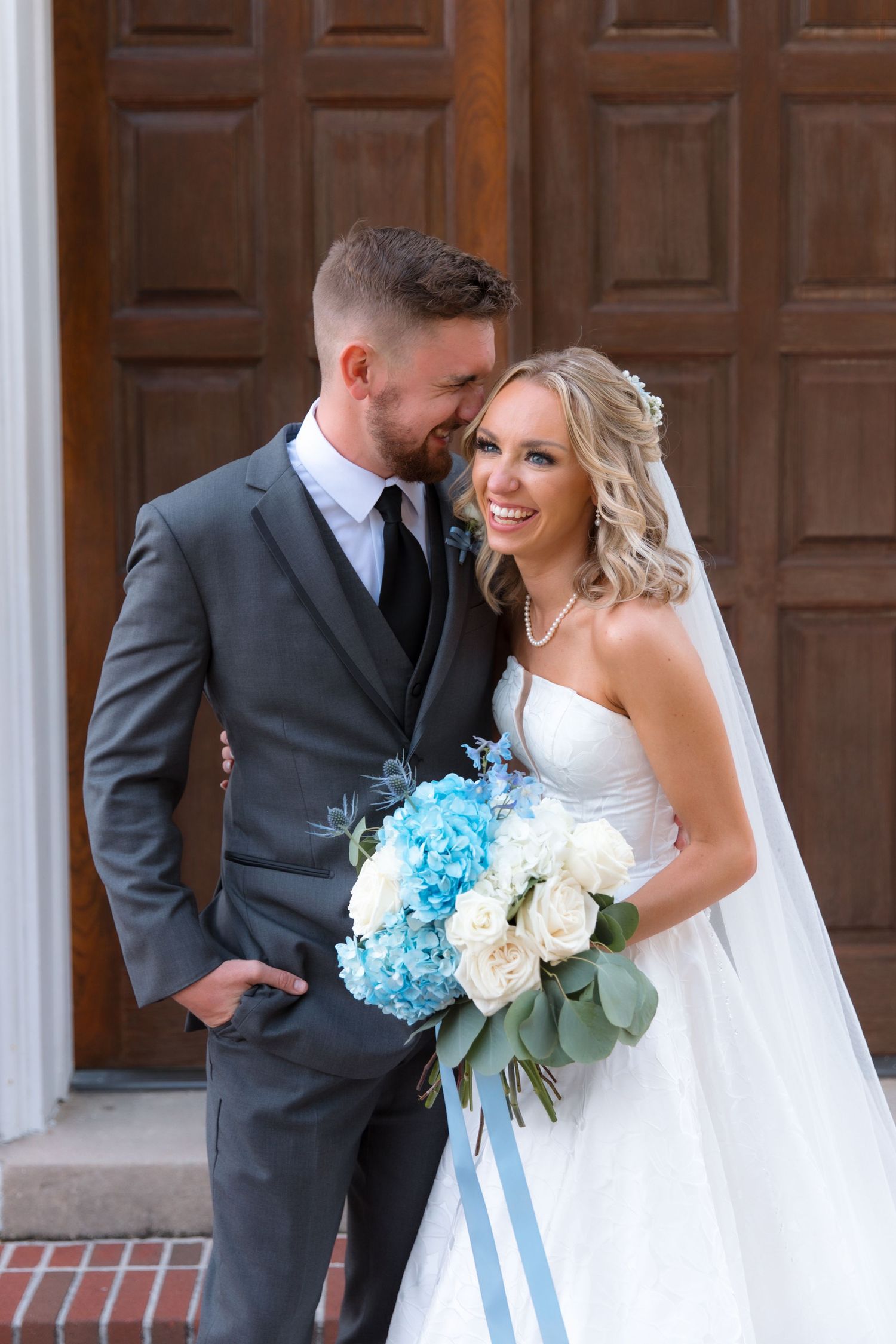 Intimate moment between couple with blue and white bouquet against wooden church doors.