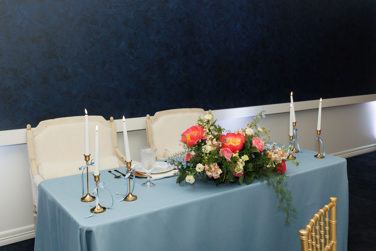 Elegant table setting with brass candlesticks and pink floral centerpiece arrangement on light blue tablecloth.