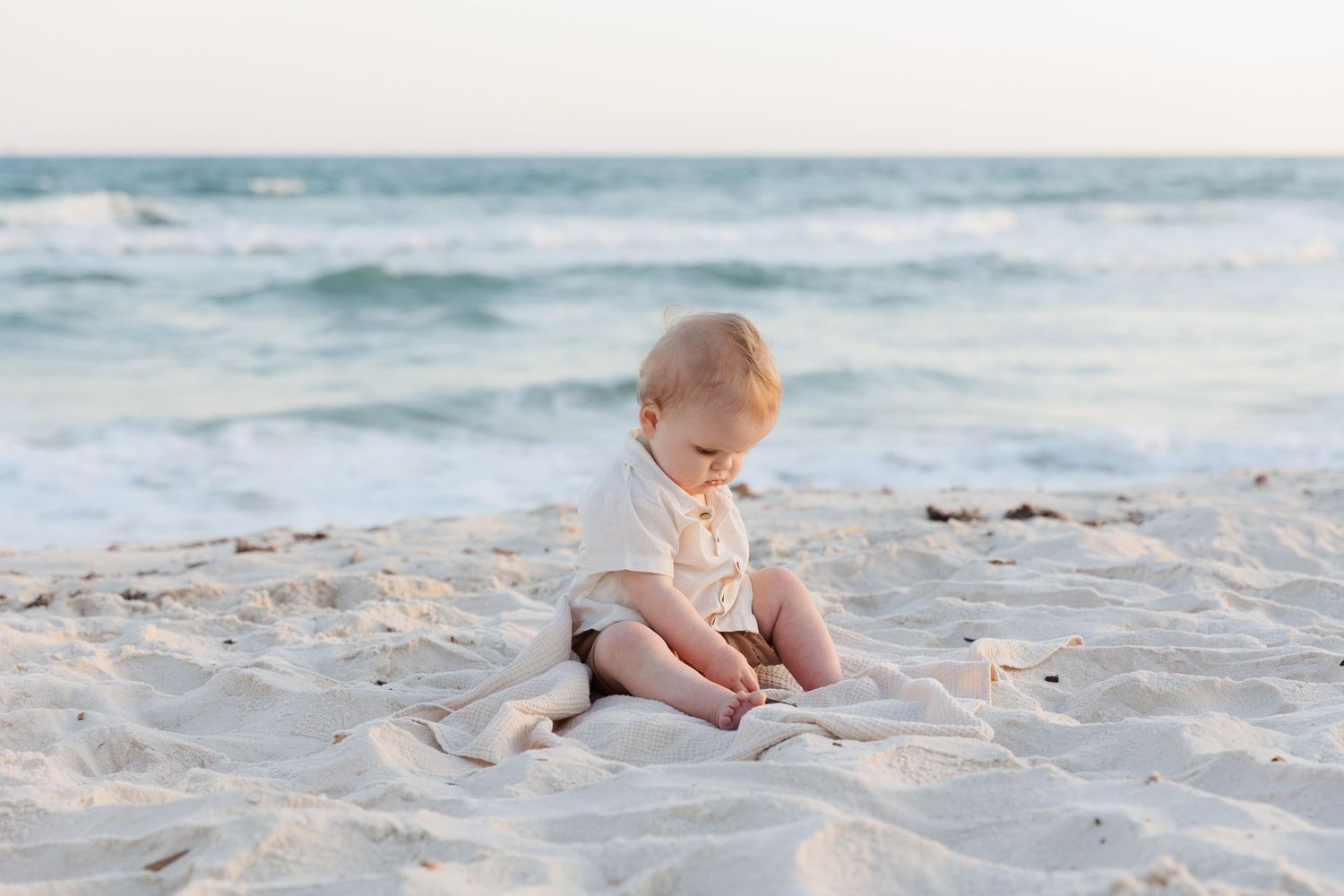 A baby sits alone on a sandy beach near ocean waves, wearing a cream outfit, looking down at the sand.