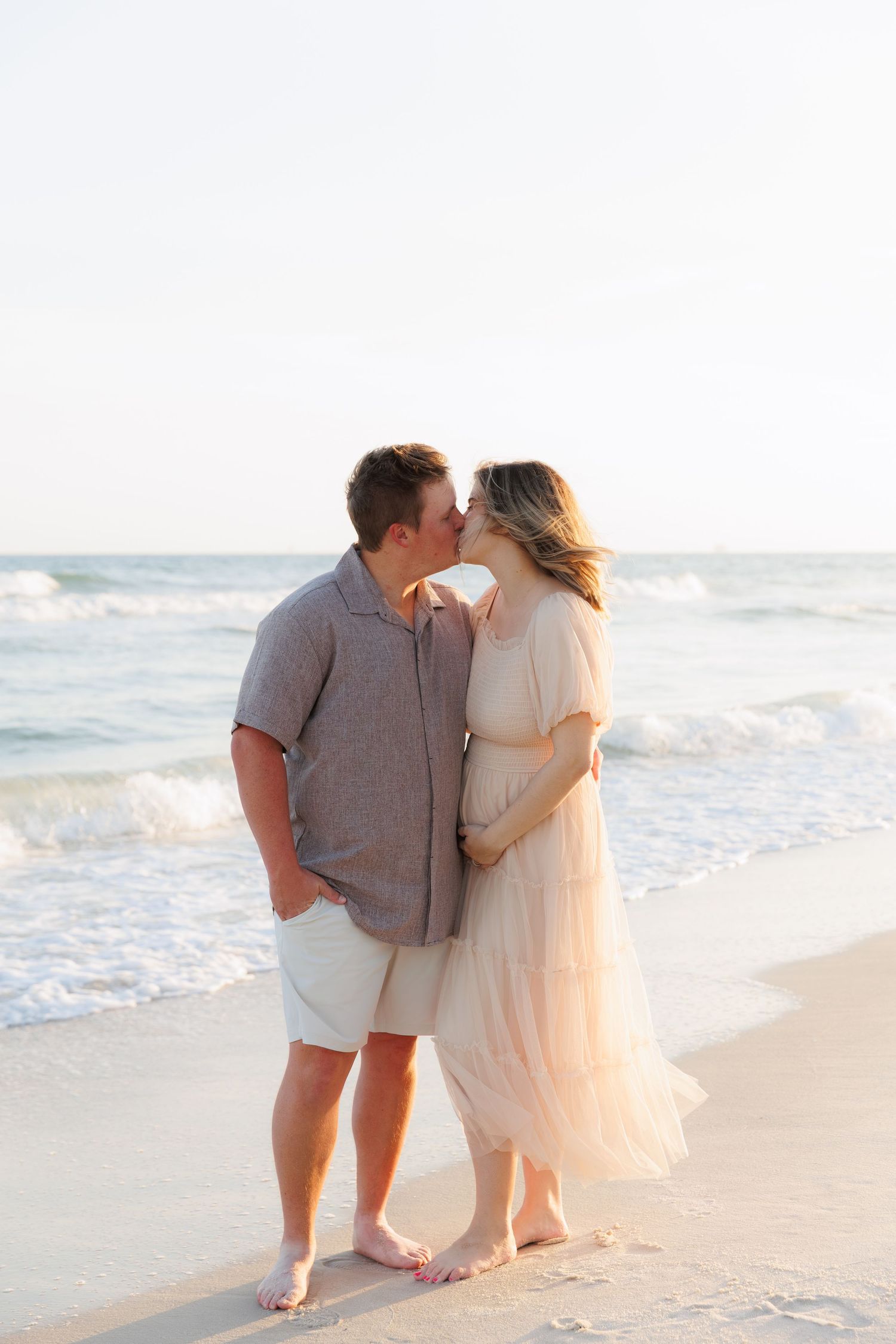Pregnant couple sharing a kiss on a sandy beach at sunset, waves gently rolling in behind them.