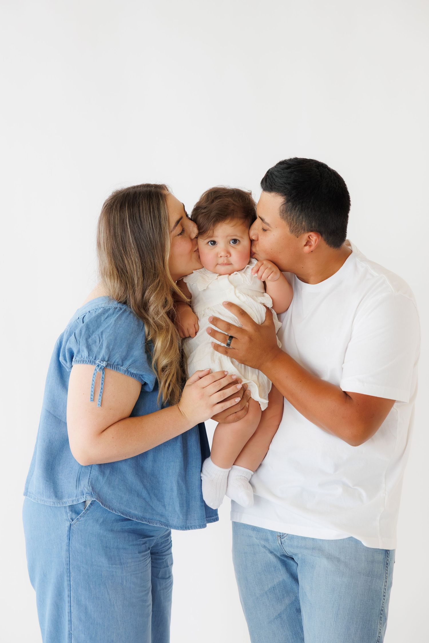 Parents kissing their baby on the cheeks in a white studio family portrait photo.