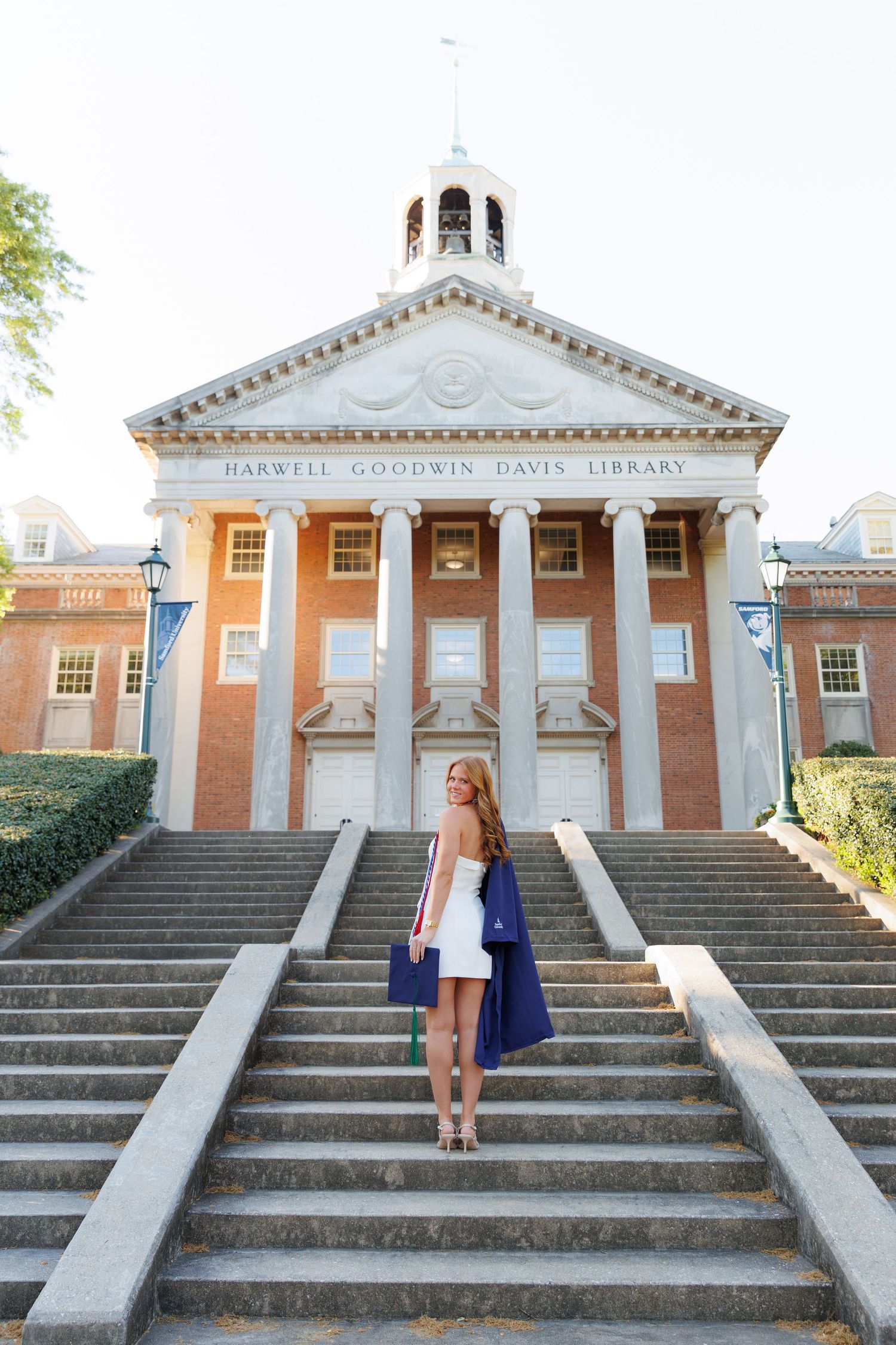 Young woman in white dress holding blue graduation gown on steps of Harwell Goodwin Davis Library.