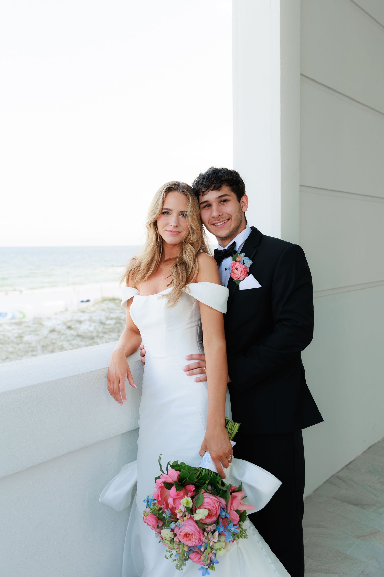 A wedding couple poses on a beachfront balcony with a colorful floral bouquet against white architecture.
