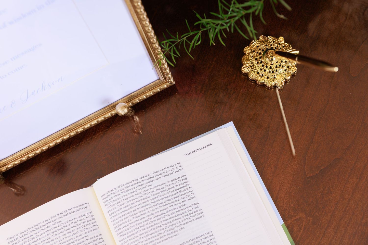An open book and framed glass item on a wooden surface with a sprig of greenery and gold glitter decoration.