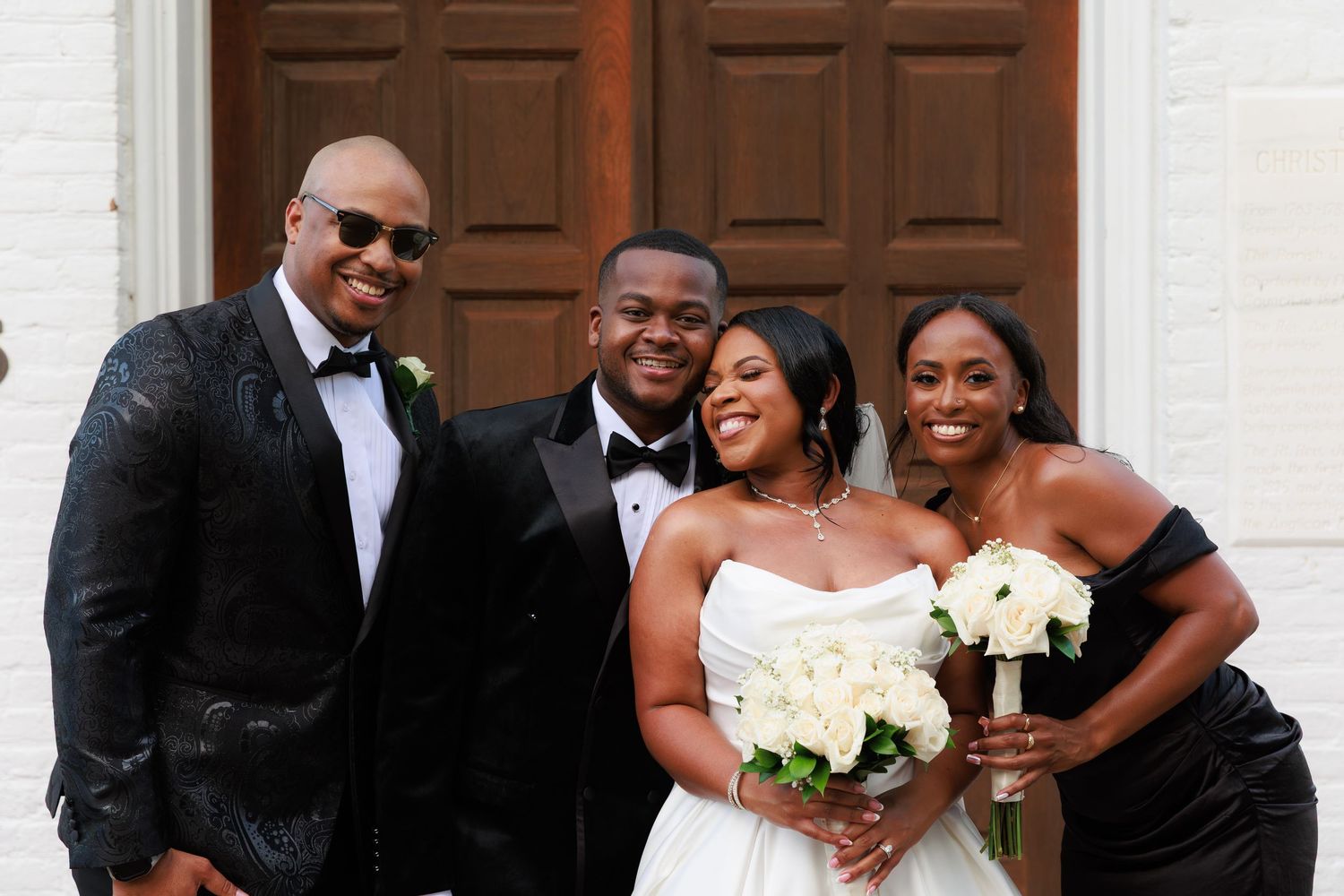 Wedding party members in formal black and white attire pose together with bouquets outside a building.