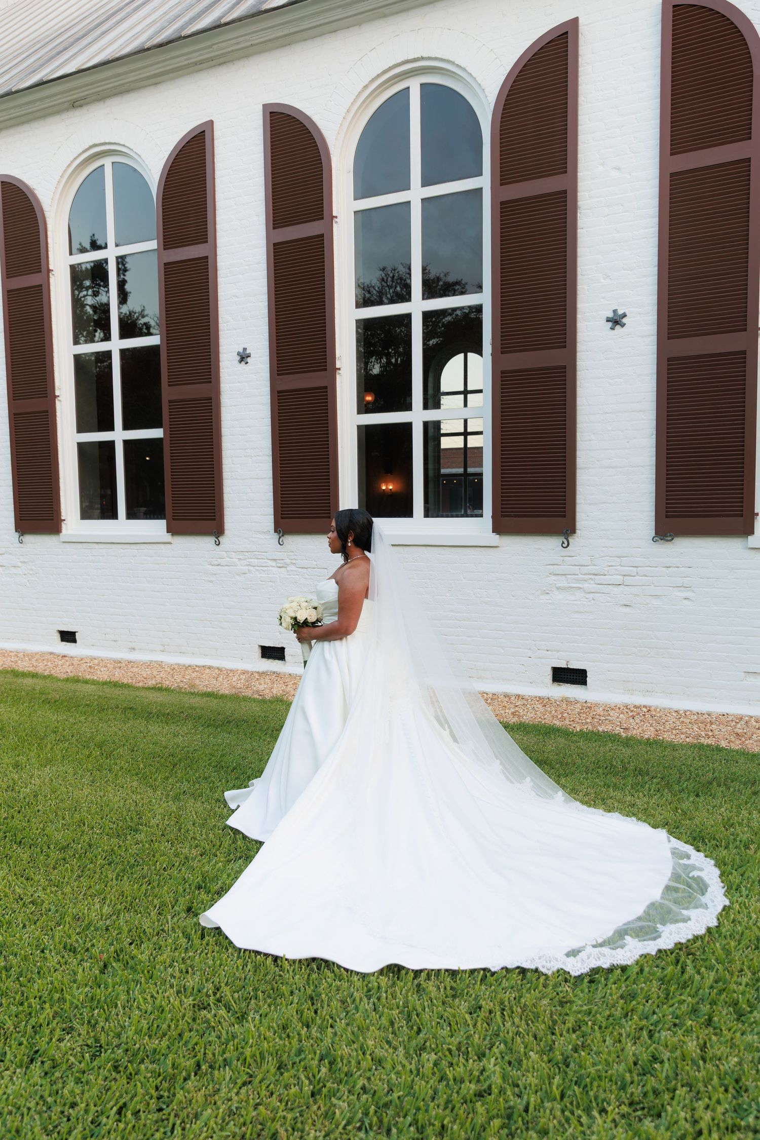 A white wedding dress train spreads elegantly across green grass in front of a white building with brown shutters.