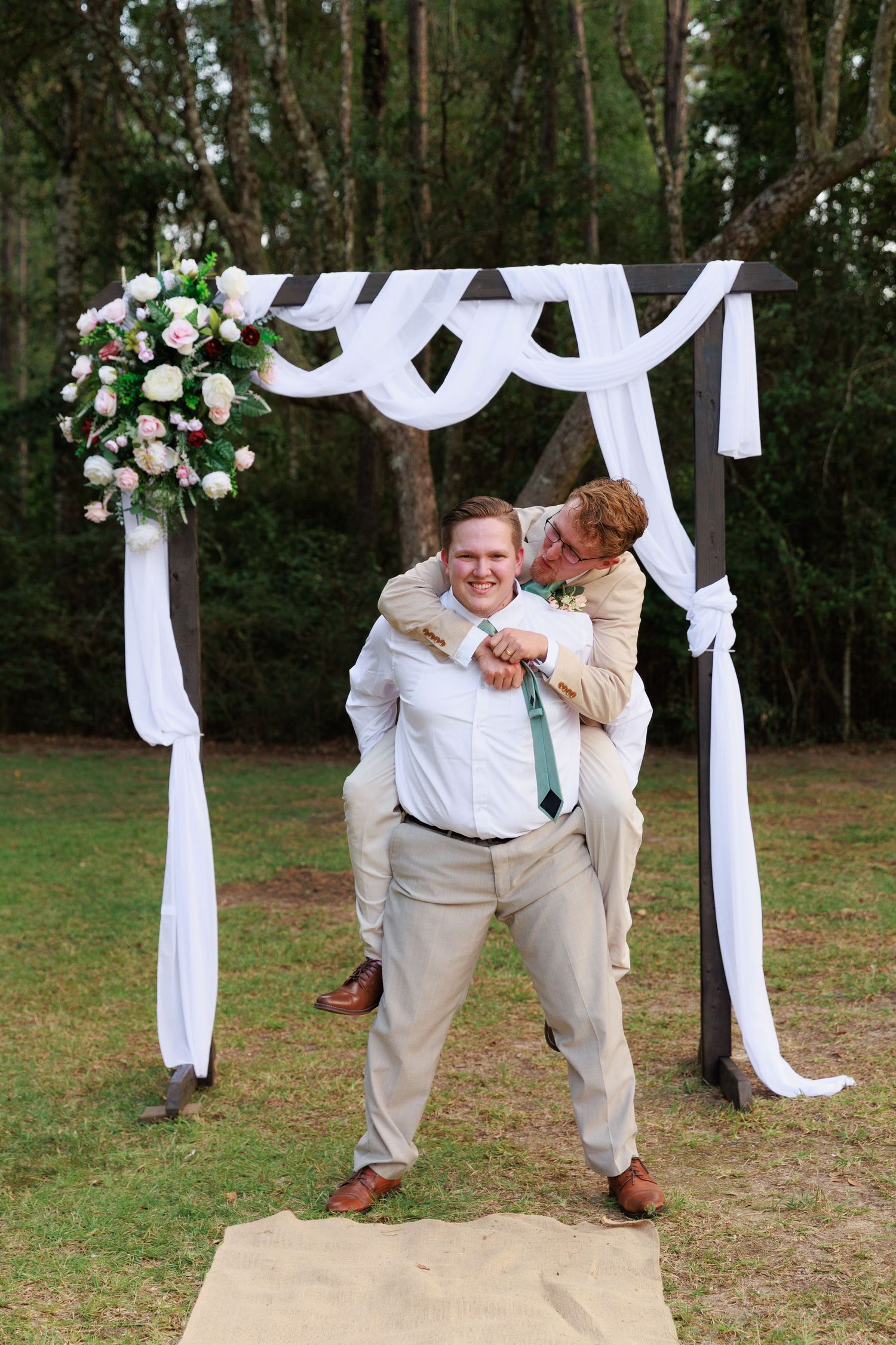 Groomsmen in khaki suits and white shirts playfully pose together under a white draped wedding arch in a wooded setting.