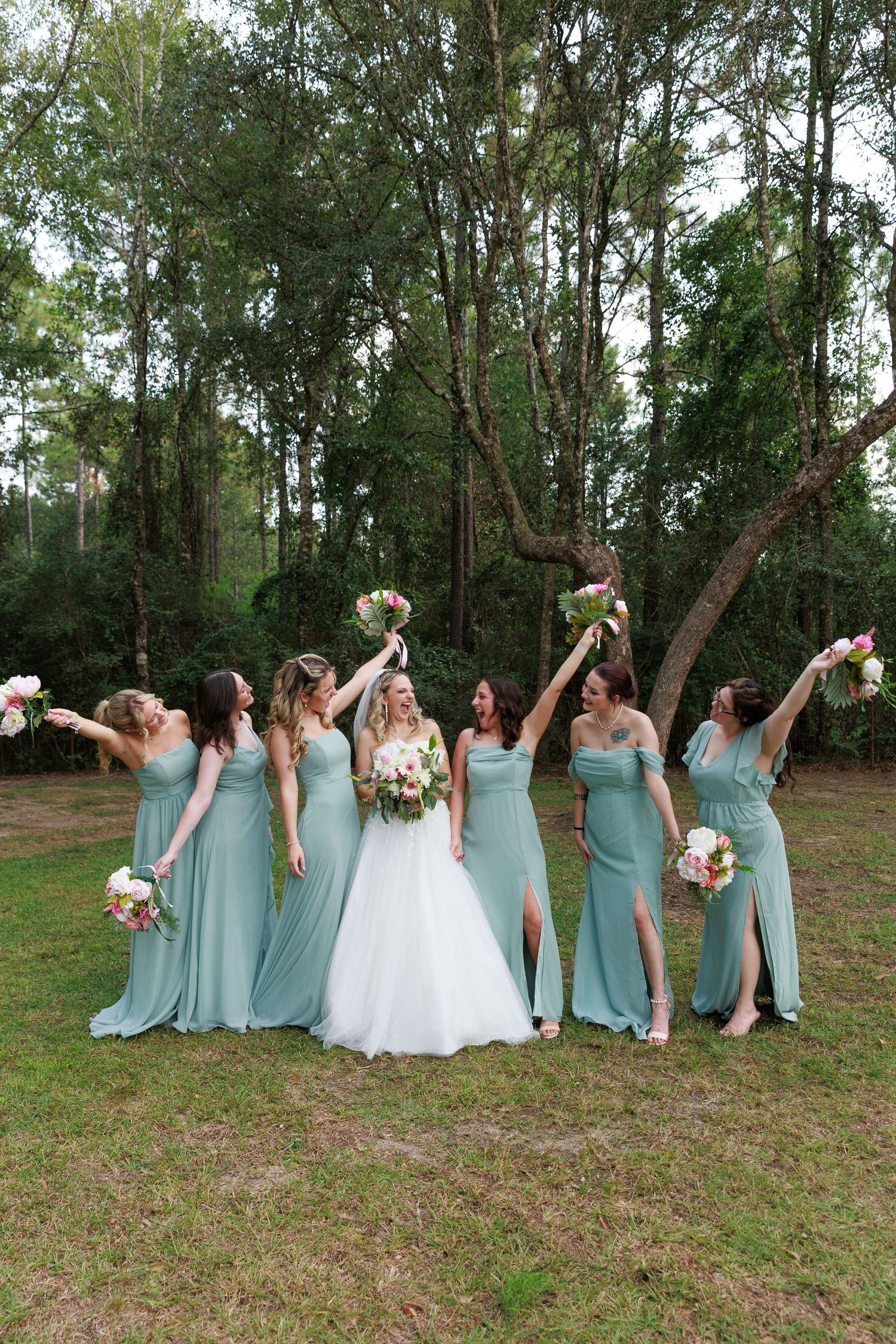 Bridal party in mint green dresses celebrates joyfully with bouquets raised in outdoor forest setting.