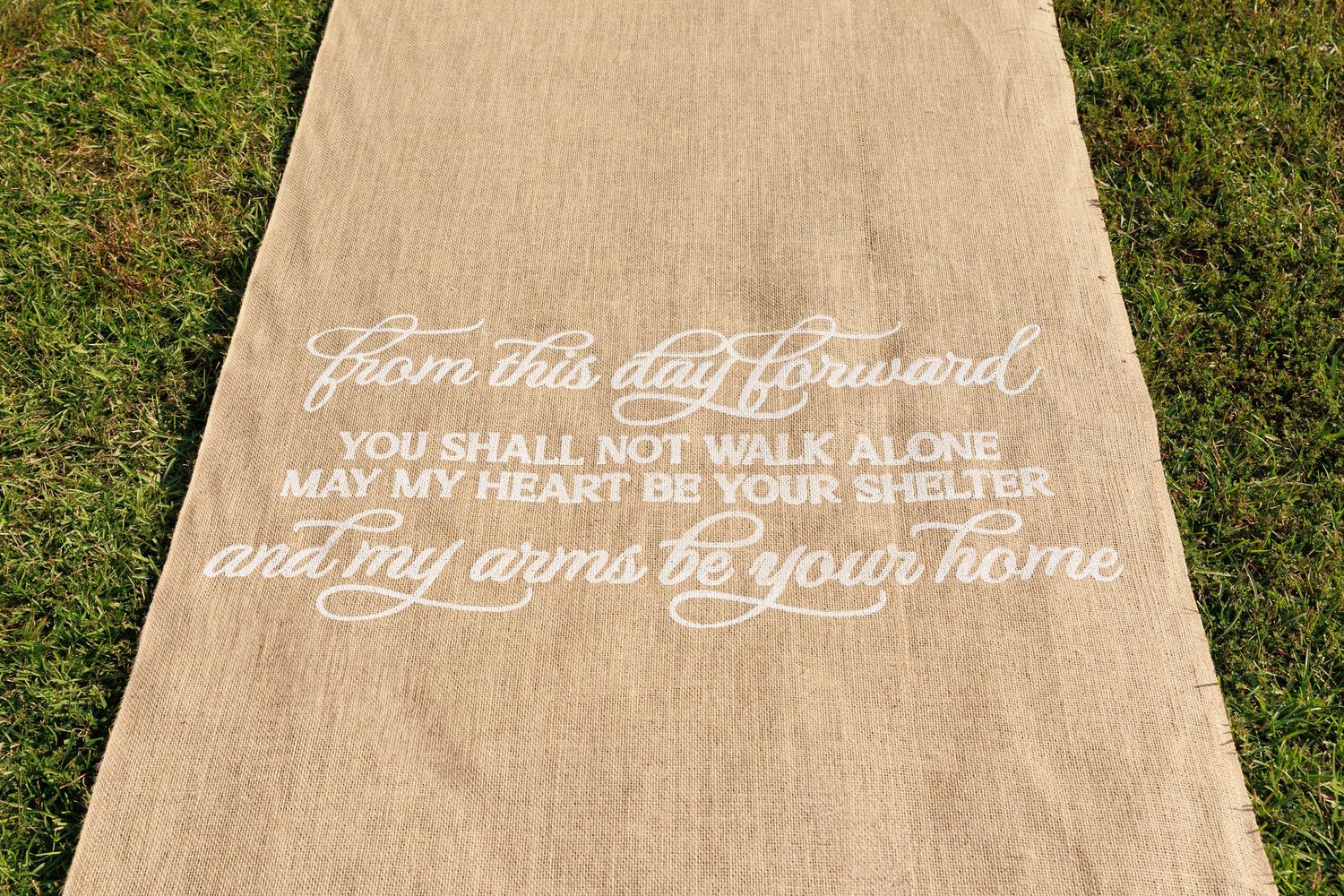 A burlap aisle runner with elegant white script text reading 'From this day forward' for a wedding ceremony.