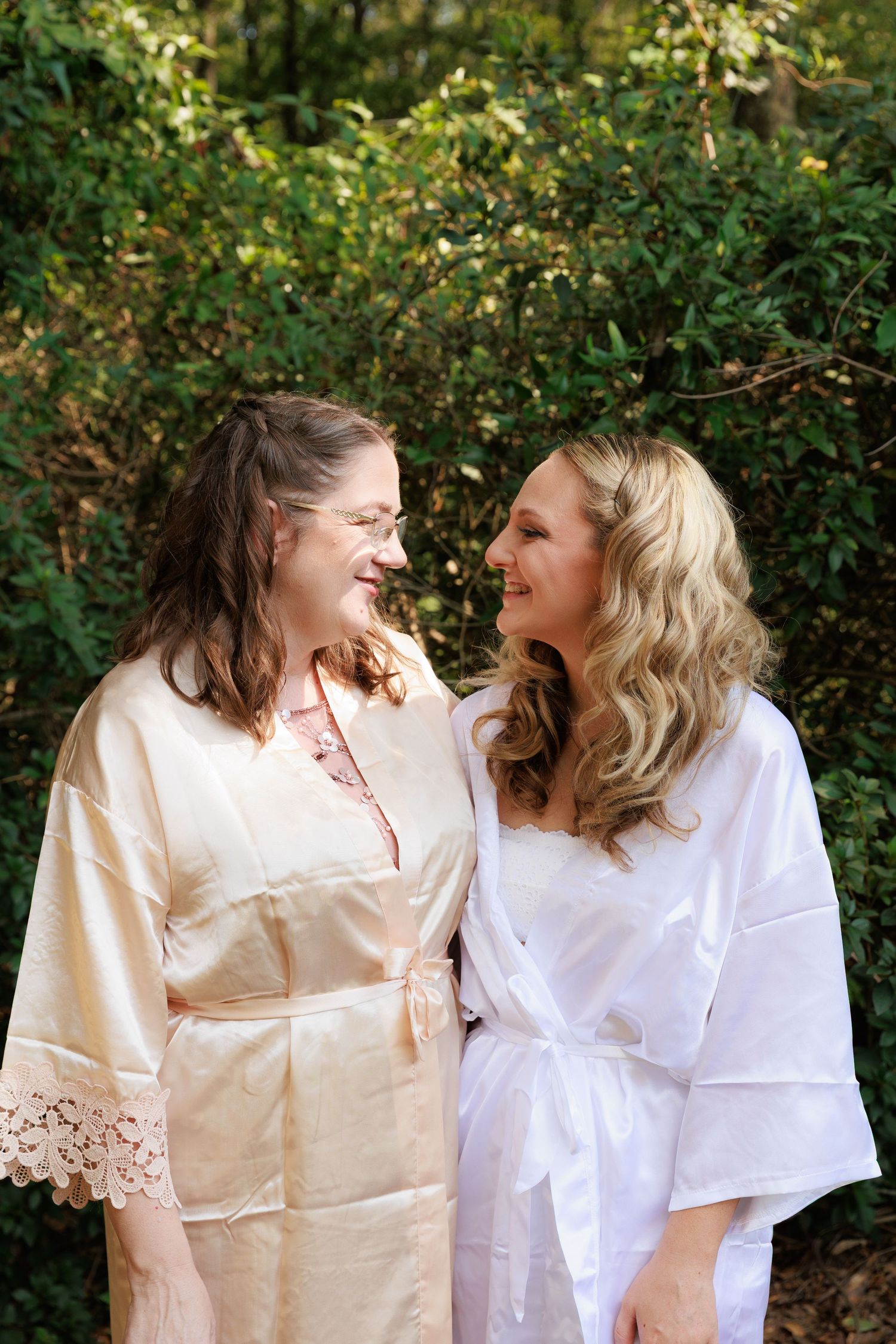 Two women in white and cream colored silk robes share a joyful moment together outdoors against green foliage.