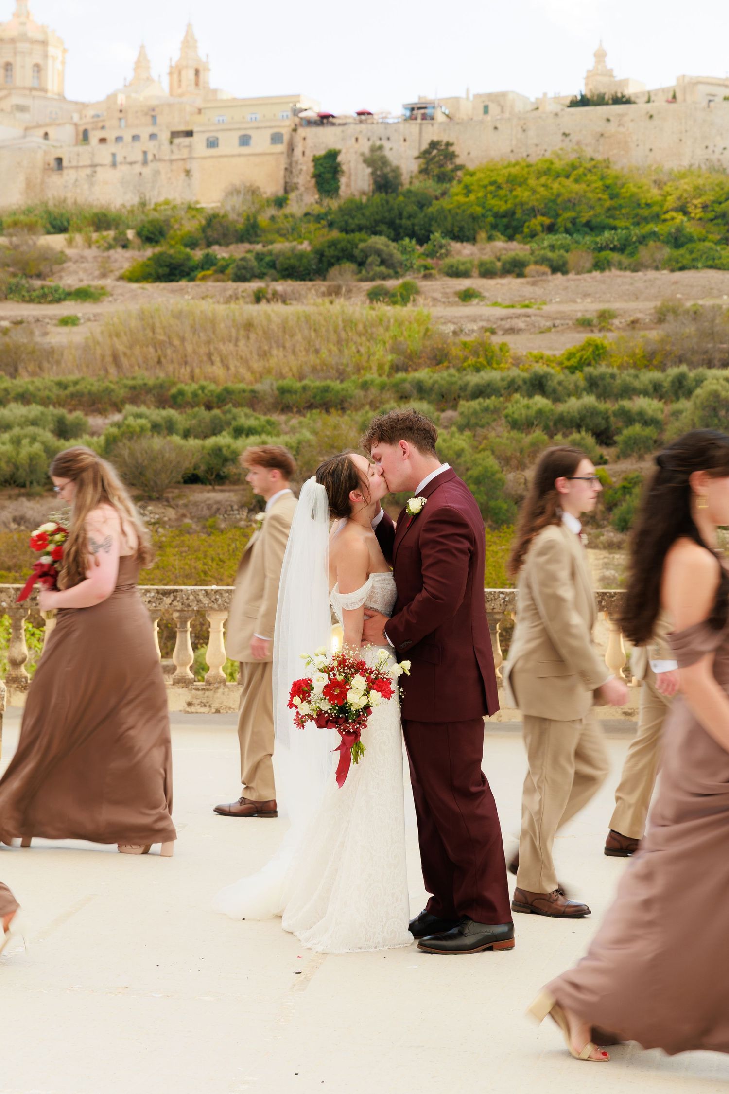 Wedding party poses for photos on sandy terrain with historic stone buildings in the background.