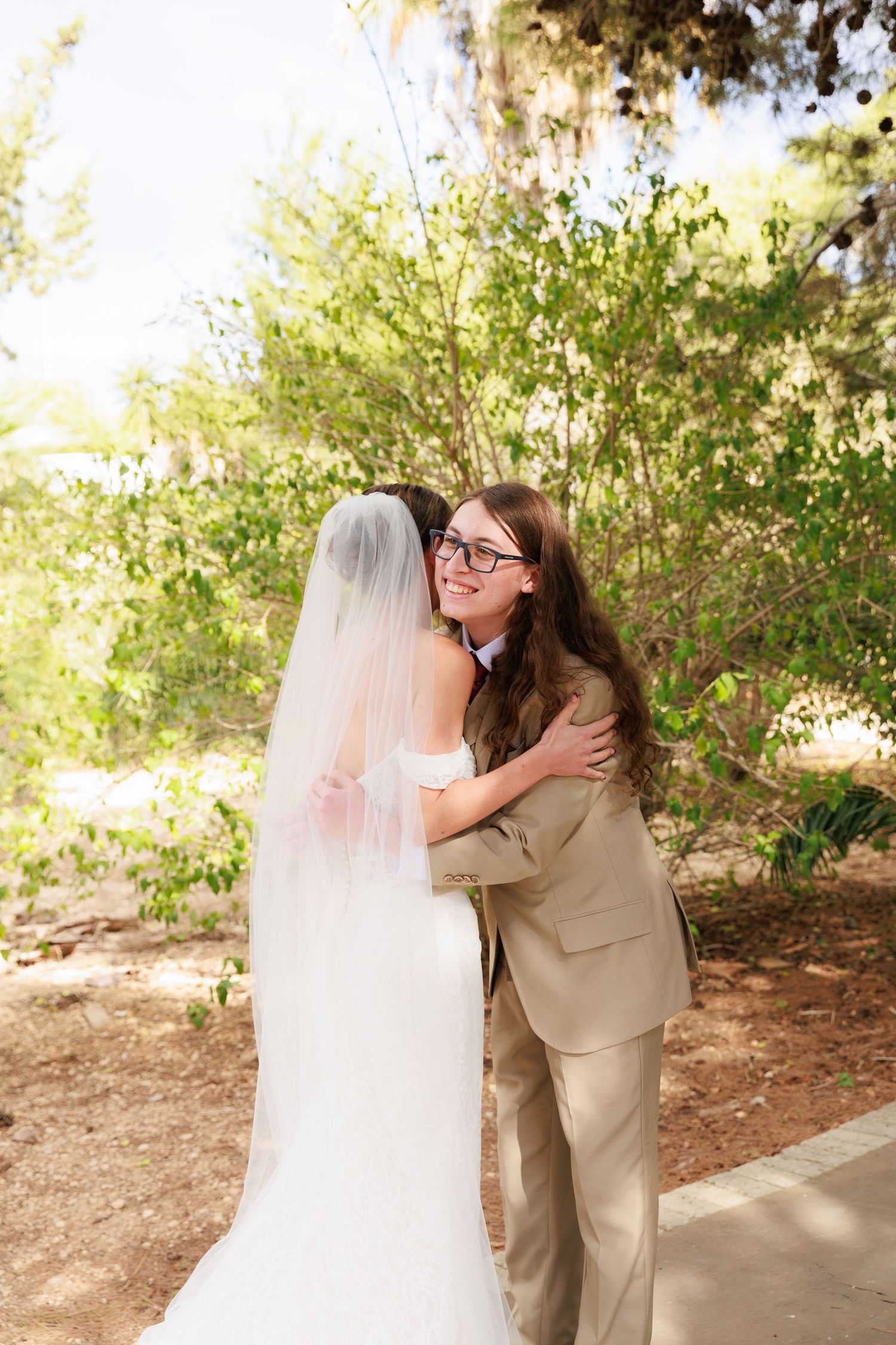 Two people share a heartfelt embrace on a garden path during a wedding ceremony, with sunlit greenery in the background.
