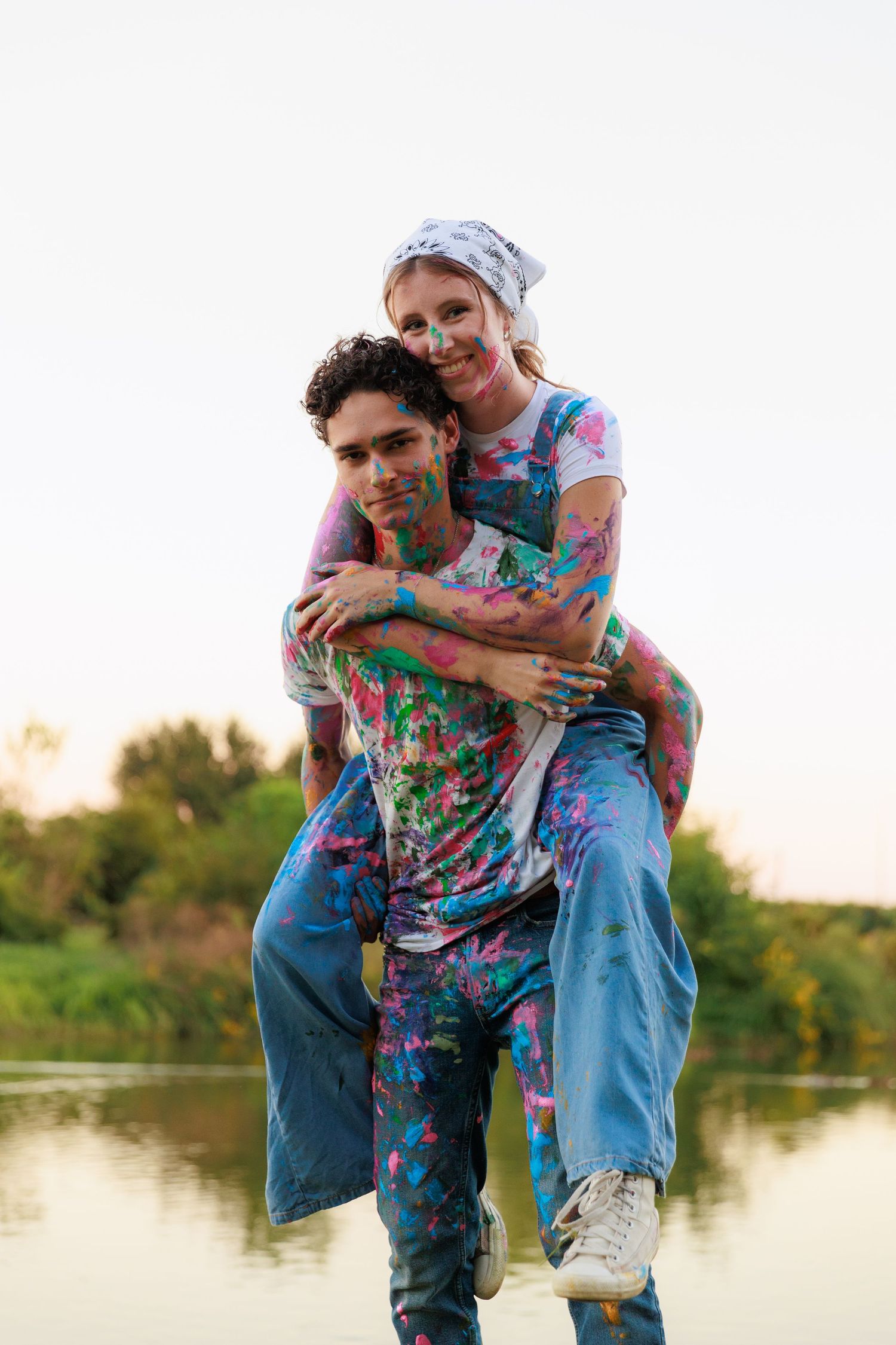 Two people covered in colorful paint embrace and laugh together by a lake at sunset.