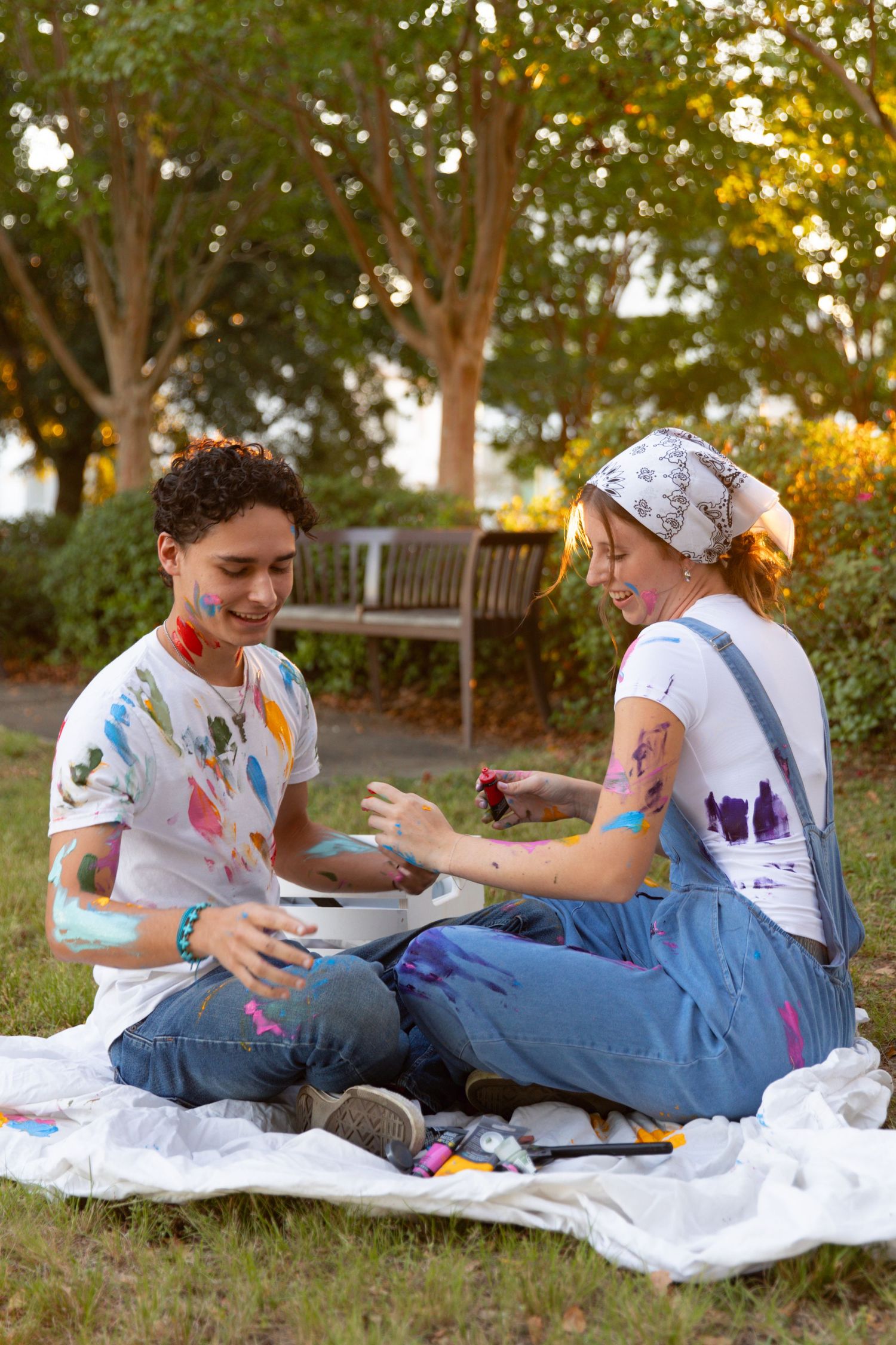 Two people sitting on a white blanket outdoors engage in a playful paint fight at sunset.
