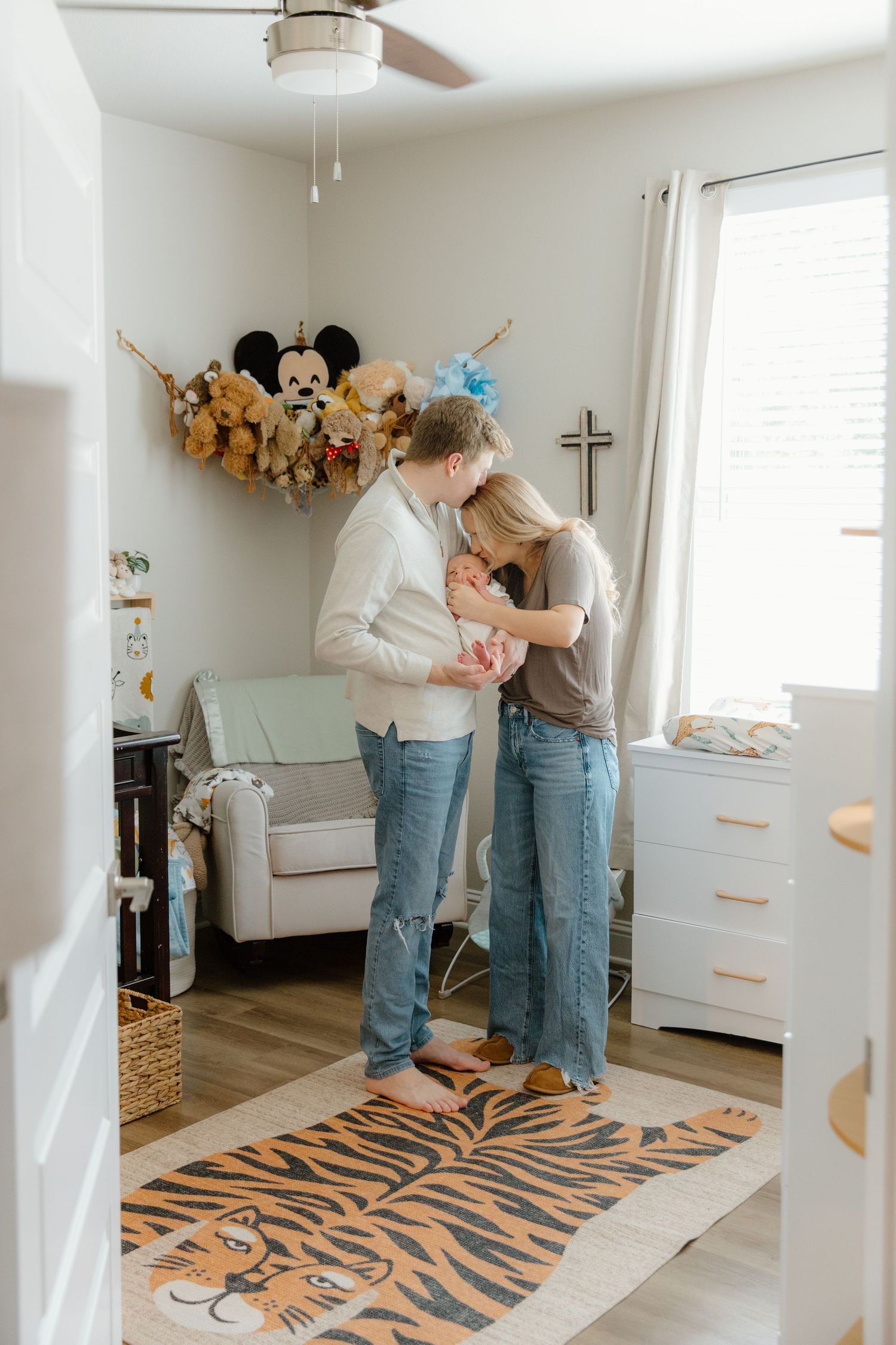 Parents share sweet moments with their baby in a bright nursery decorated with stuffed animals and a tiger print rug.