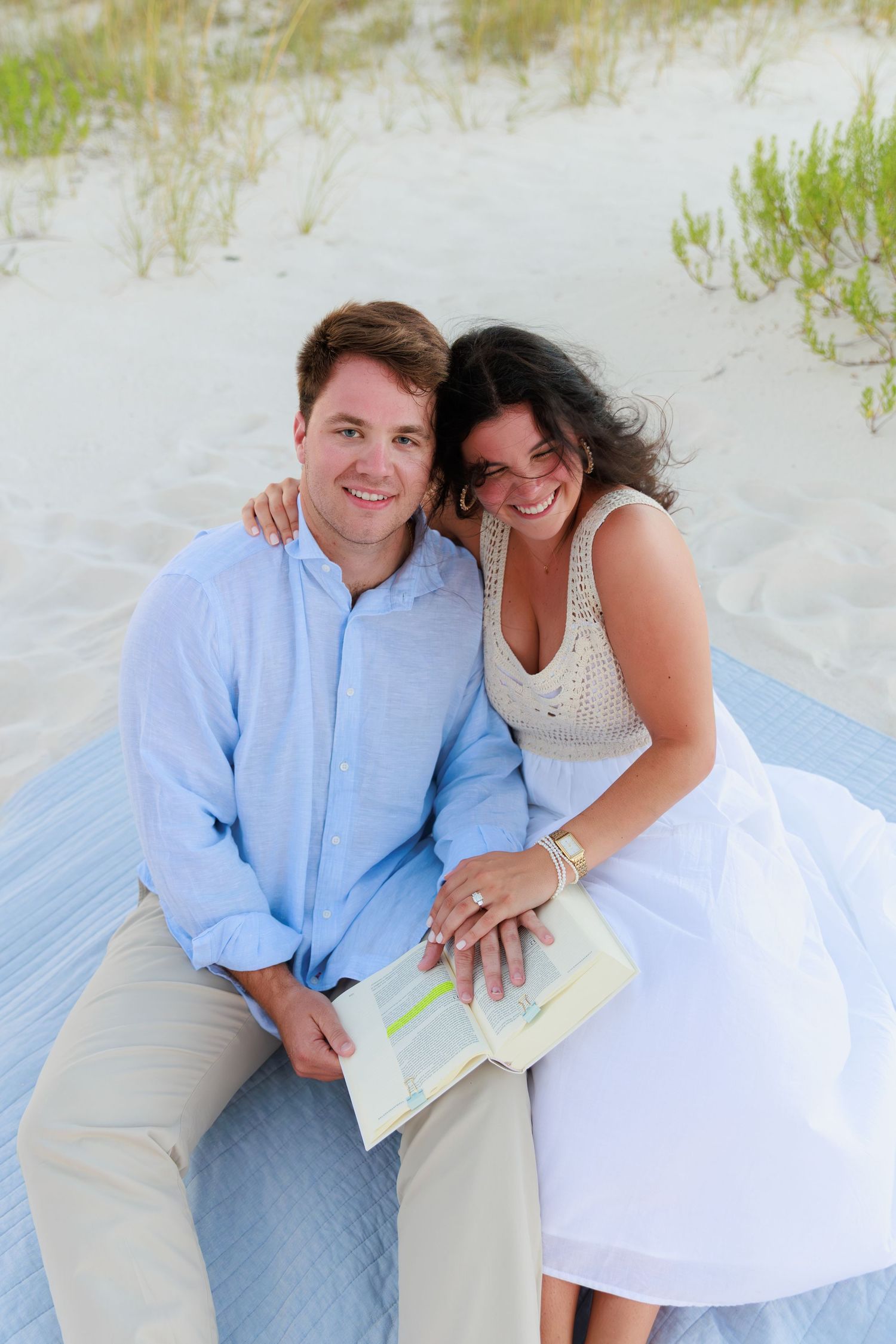 A couple sits together on a blue blanket at the beach with sand dunes and sea grass in the background.