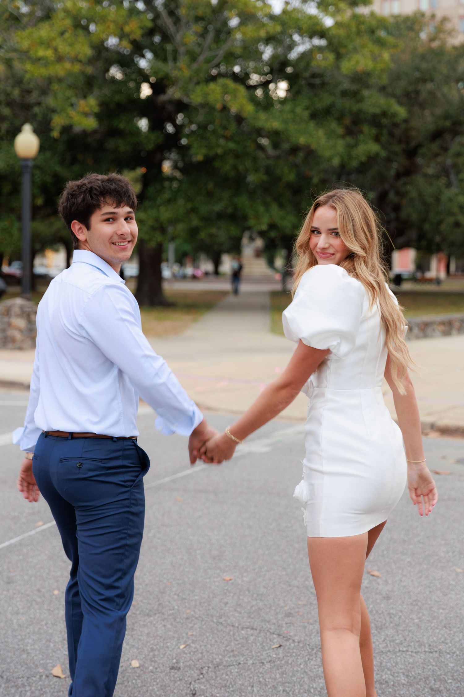 A couple in white and navy outfits playfully dances together on a paved path surrounded by green trees.