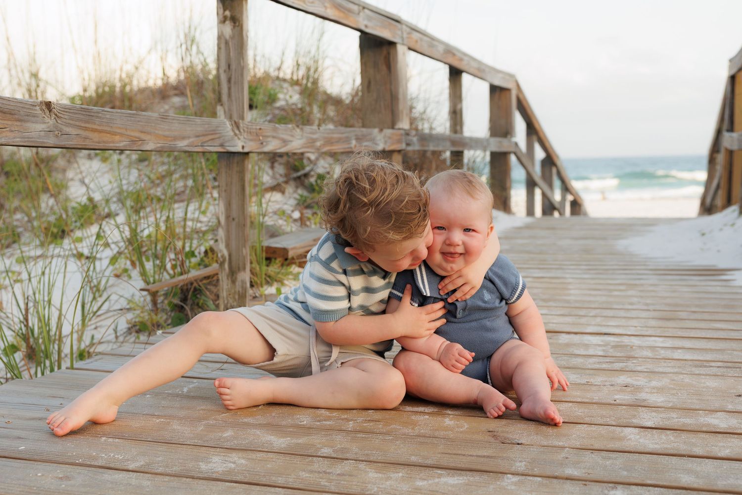 Two young boys laughing and hugging on a wooden beach boardwalk with ocean views.