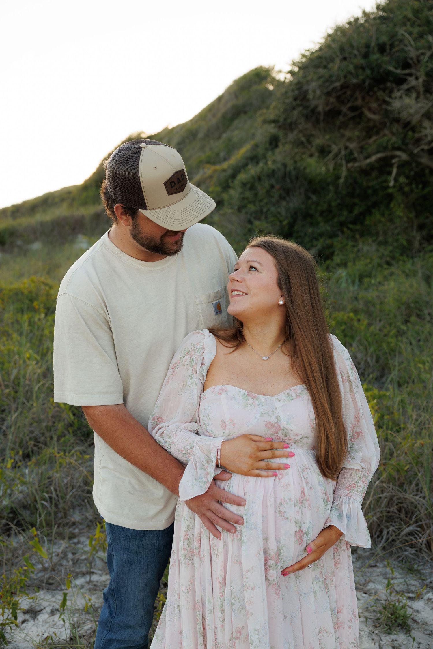Couple sharing a tender moment on sand dunes, pregnant woman in floral dress cradling baby bump at sunset.