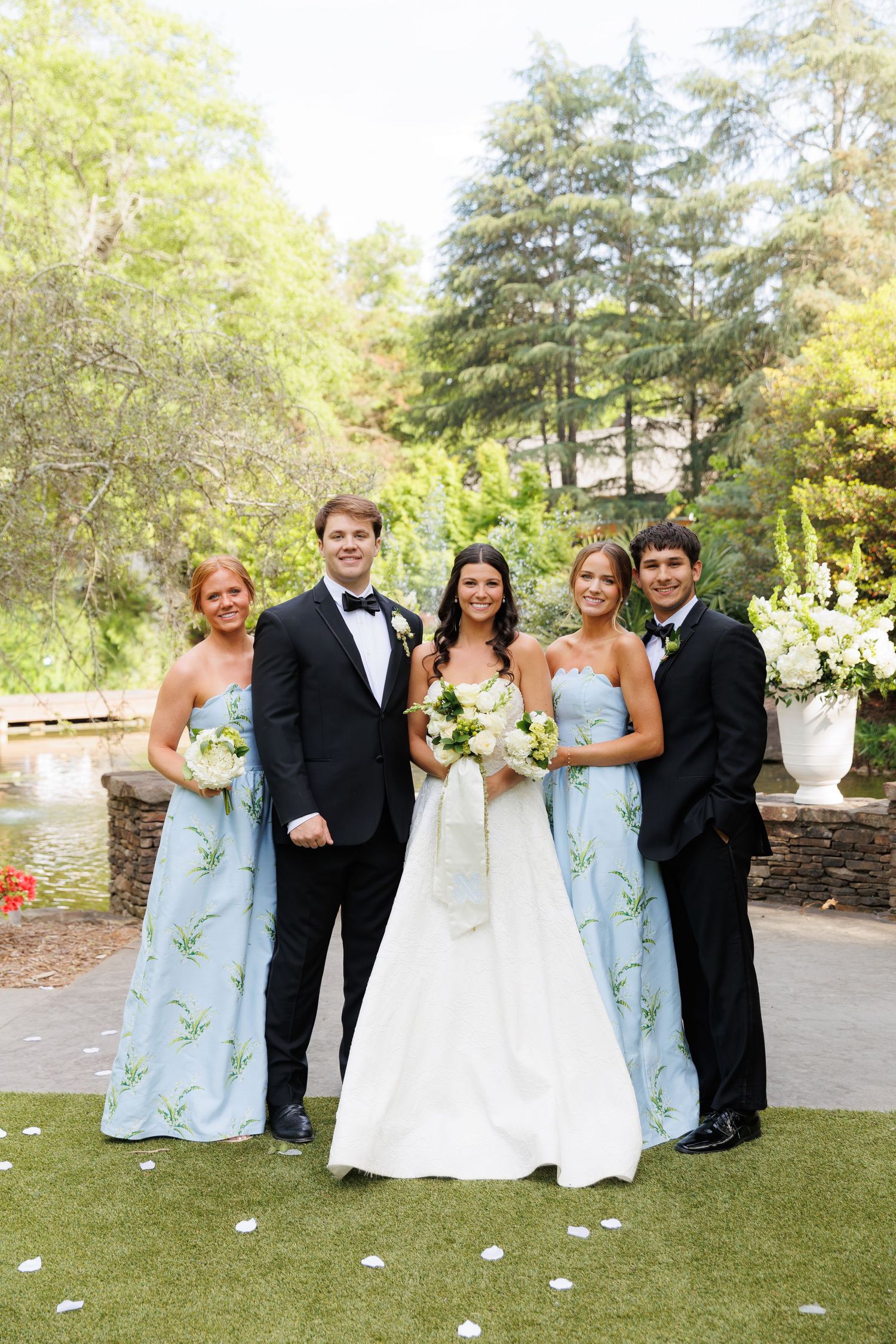 Bride and groom pose with bridesmaids in light blue gowns and groomsman in tuxedo at outdoor wedding venue.