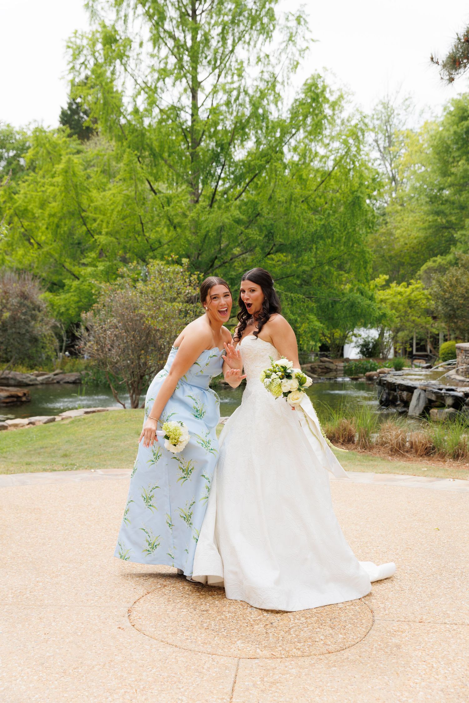 Bride in white gown and bridesmaid in blue floral dress pose with bouquets in an outdoor garden setting.