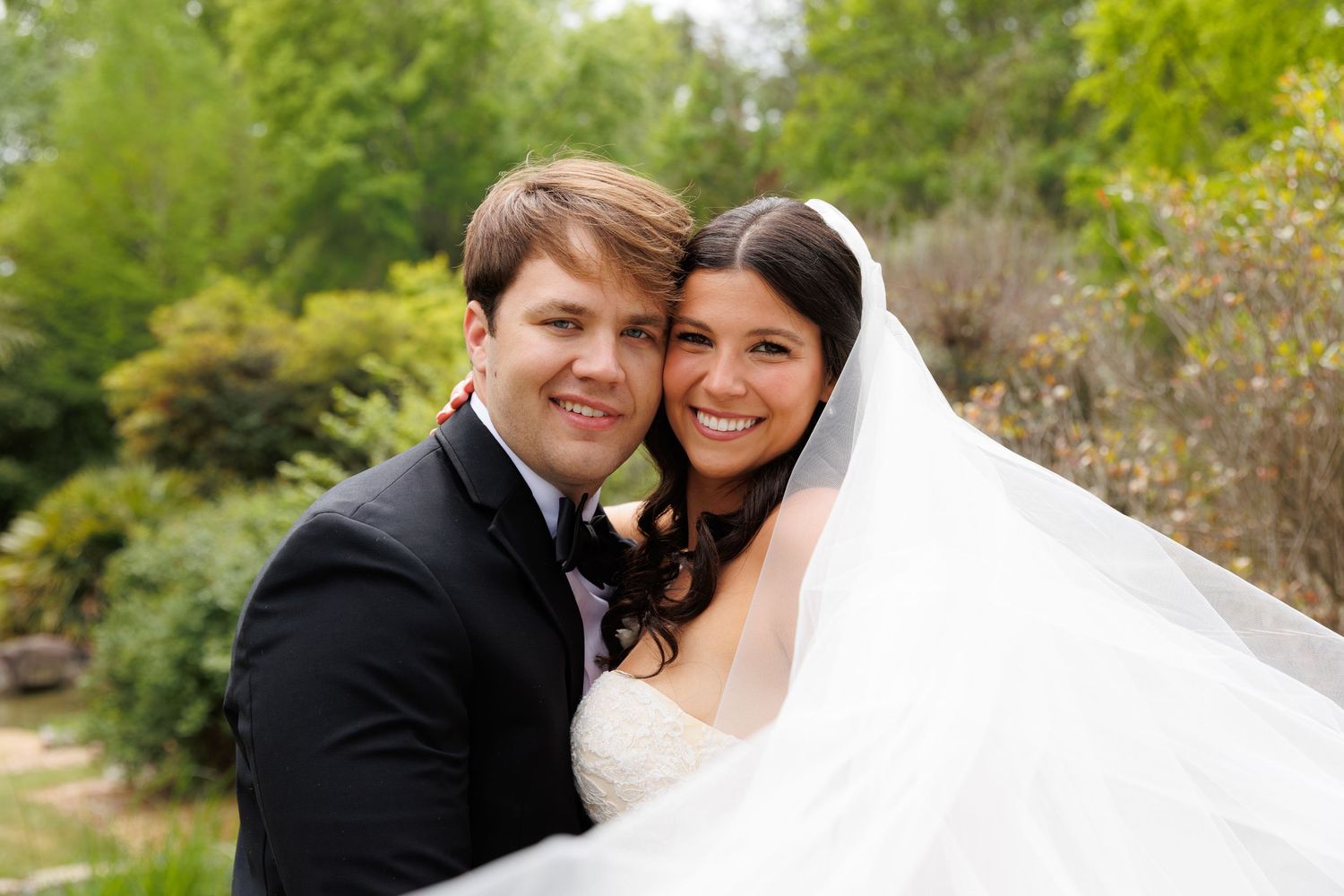 Happy bride and groom embracing outdoors in garden, bride wearing white gown and veil.