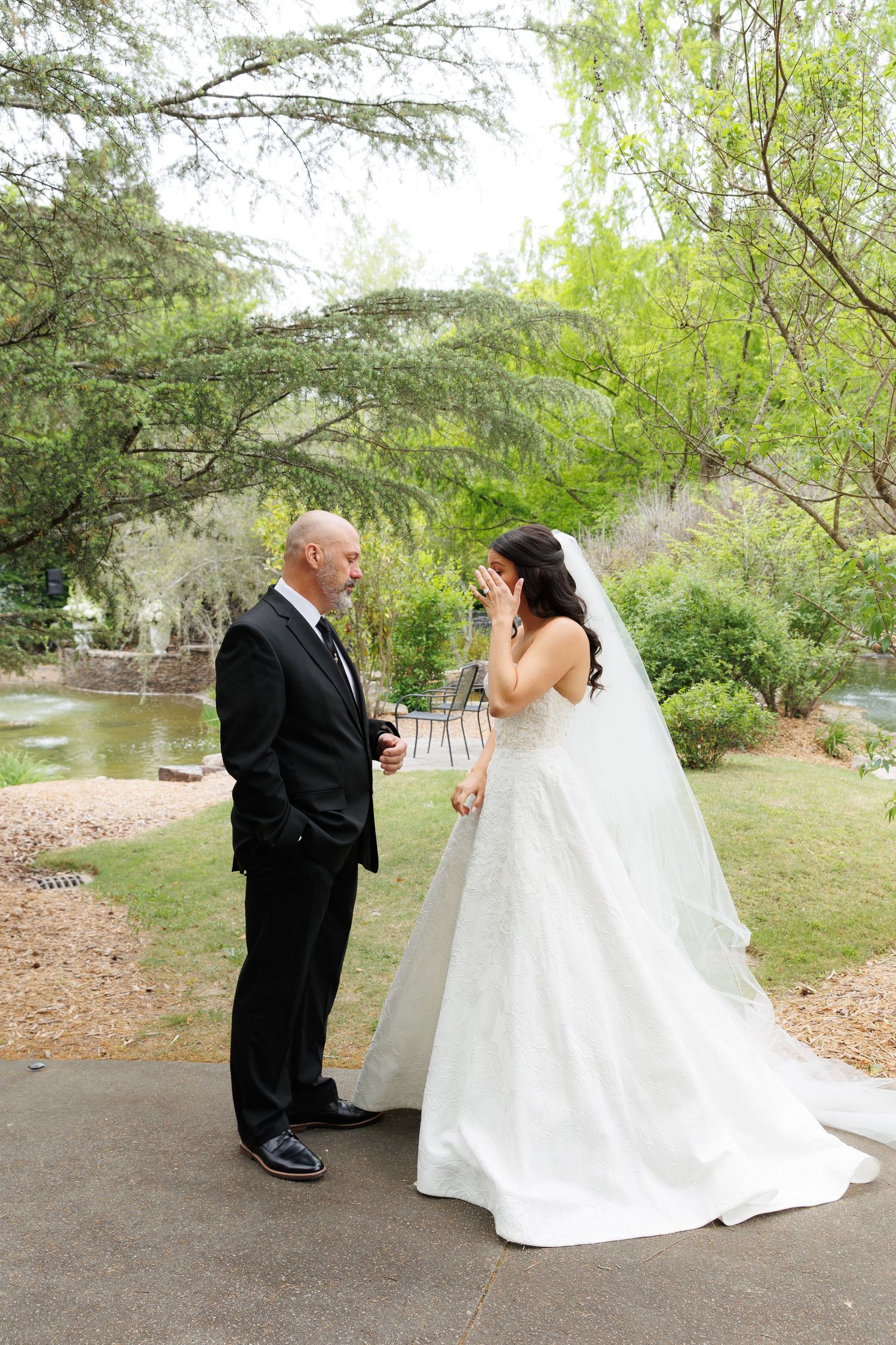 Emotional bride covering her face during first look with groom in black suit at outdoor garden wedding venue.