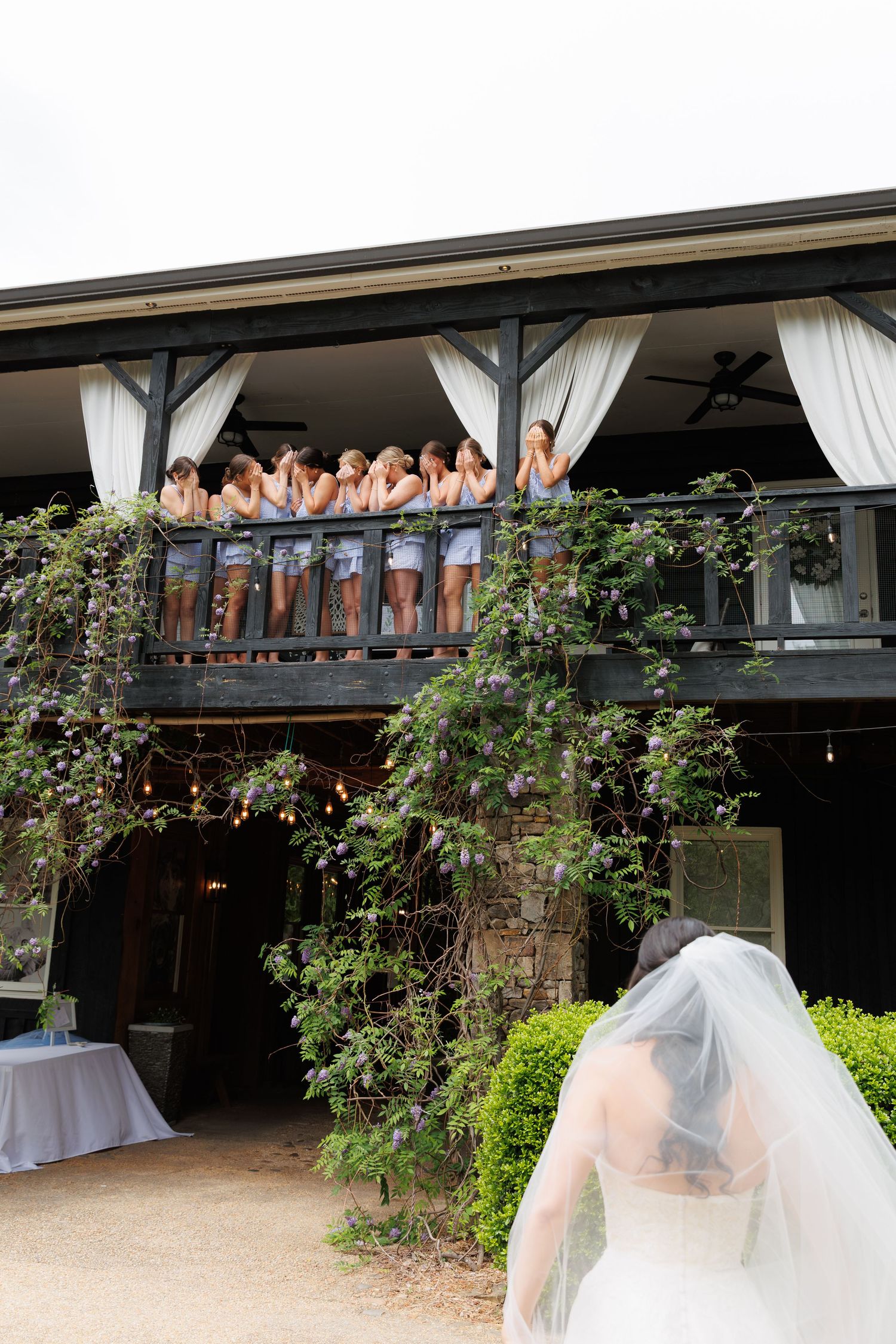 Bride in white gown and veil stands before venue balcony where bridesmaids in blue dresses react with excitement.