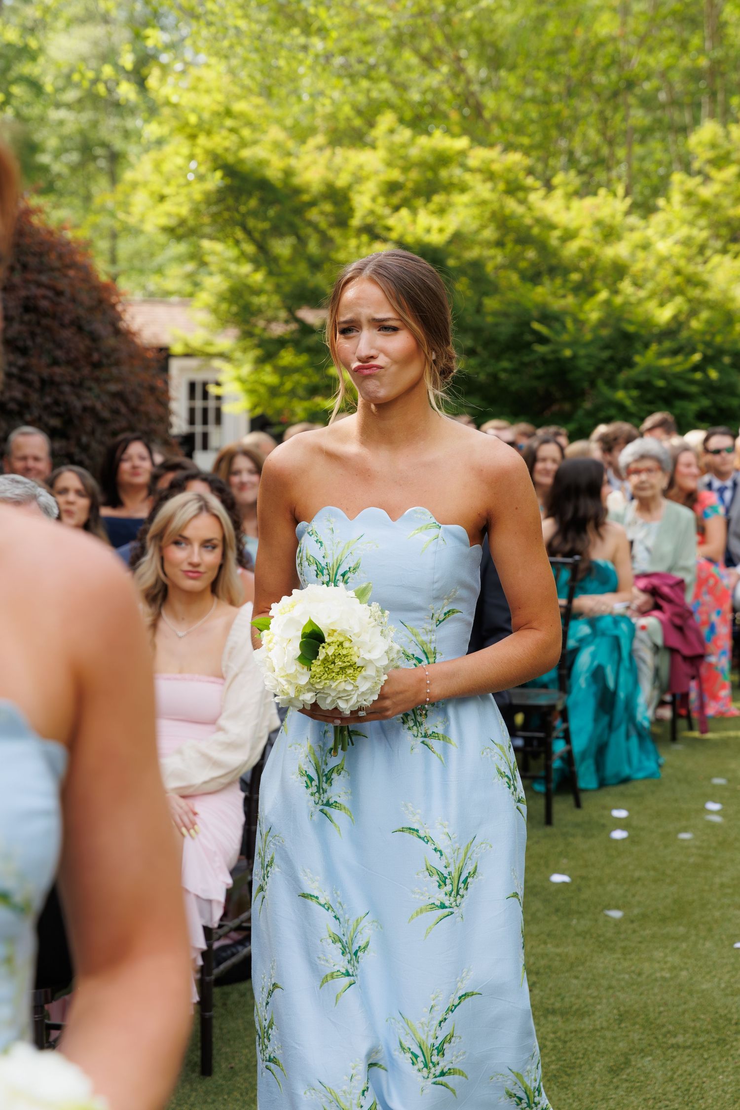 A bridesmaid in a light blue floral strapless gown holds a white bouquet while walking down the aisle at an outdoor wedding.