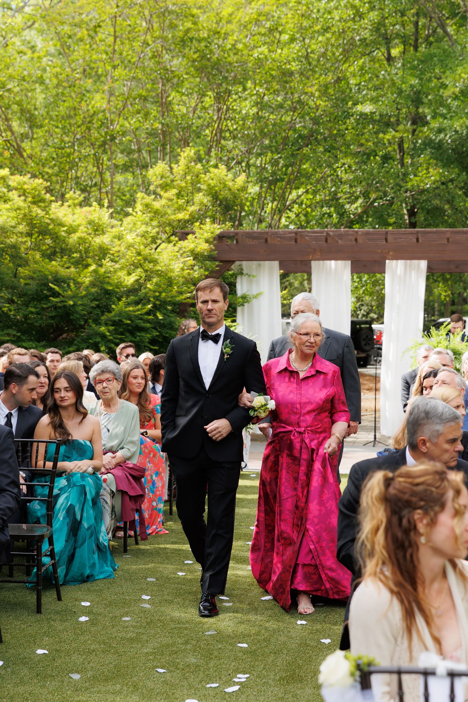 Groom in tuxedo escorts elderly woman in pink dress down outdoor wedding aisle lined with seated guests.
