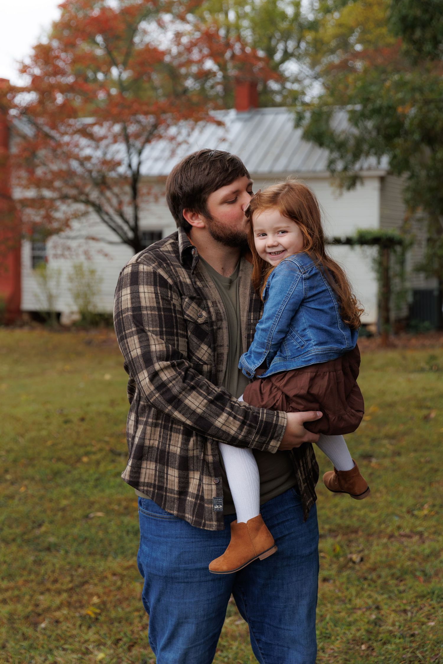 A sweet autumn moment captured outdoors with fall foliage and a white house in the background.