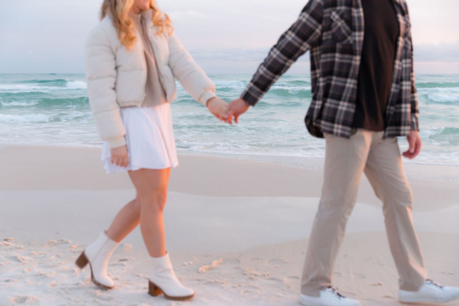 A couple strolls along a serene beach with white sand and turquoise waves in the background.
