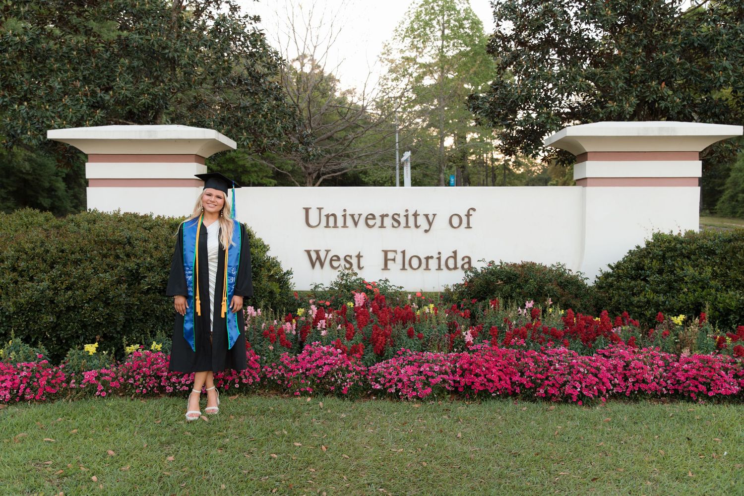 A graduation photoshoot at the University of West Florida entrance sign with vibrant pink azalea bushes in bloom.
