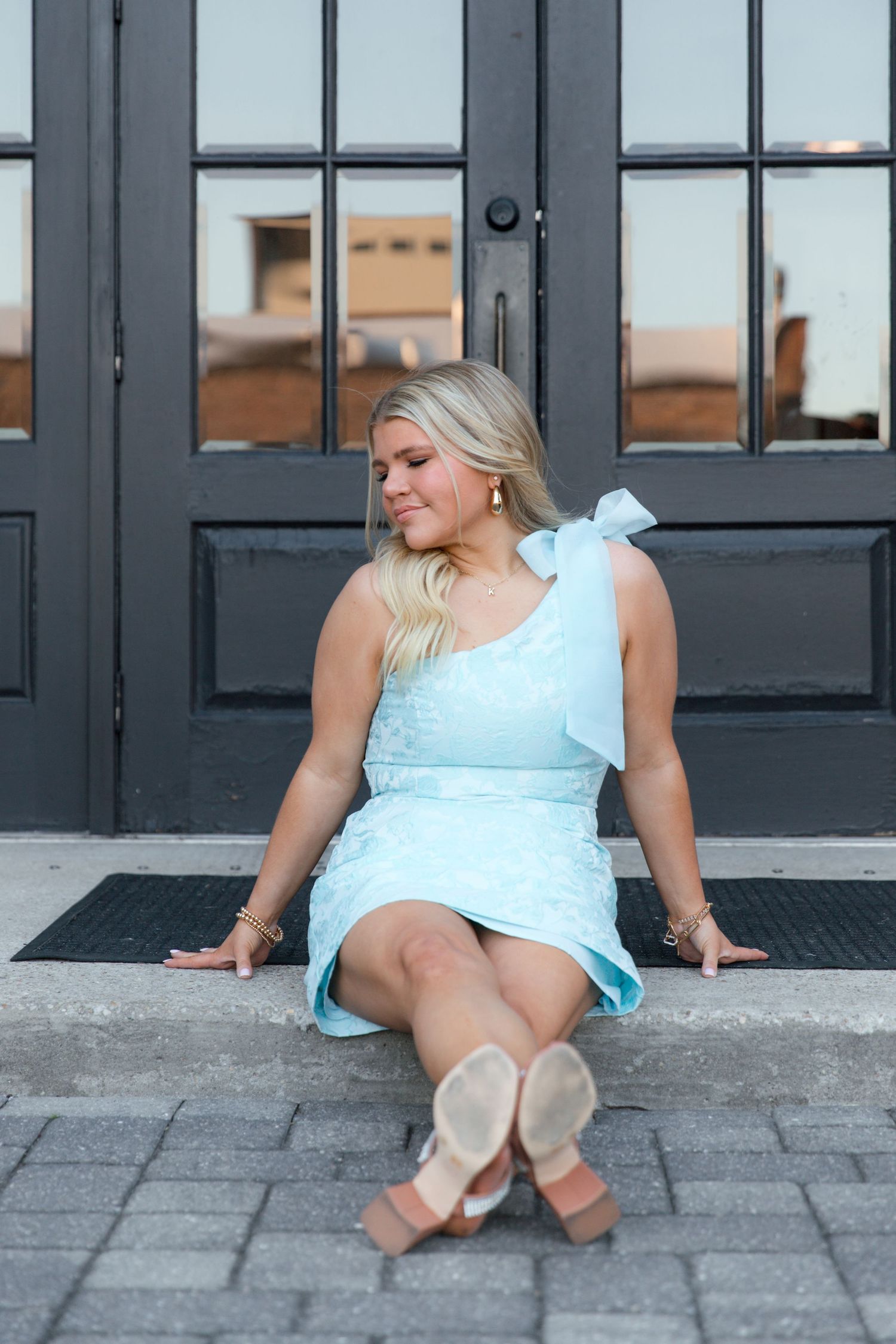 A person in a light blue dress with bow detail sits on the ground in front of black French doors.
